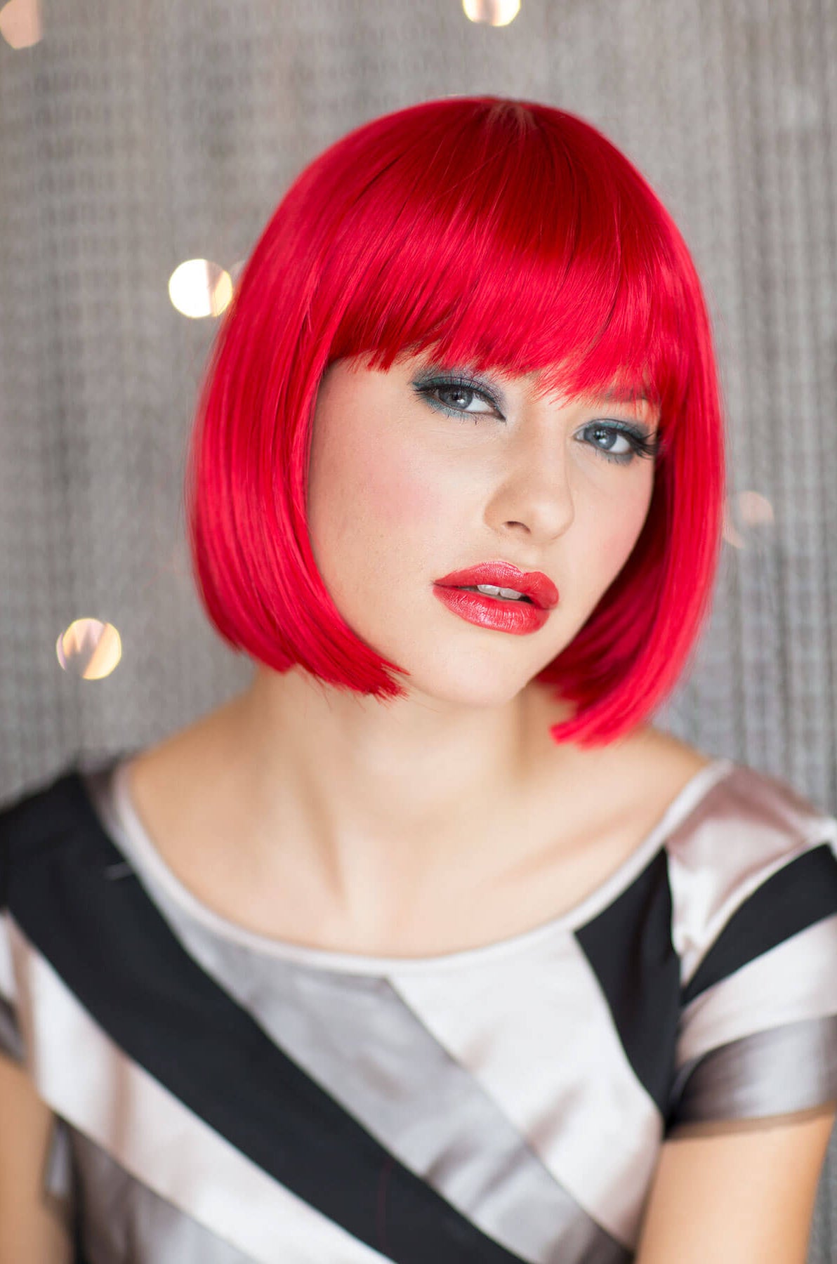 Person with bright red bob haircut and makeup against a neutral background. Vibrant, bright red bob wig: Maisy
