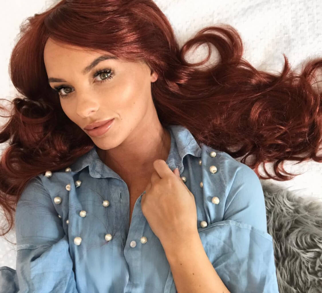 Woman with red hair wearing a blue denim shirt with pearl buttons on a white background. Long copper red wig with side sweeping fringe: Marcia