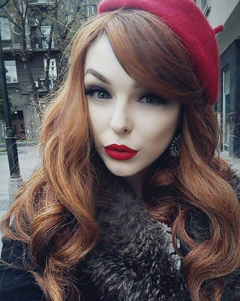 A woman with long wavy auburn hair wearing a red beret, red lipstick, and a dark fur-trimmed coat. Red & auburn wig with blonde highlights, long: Freya