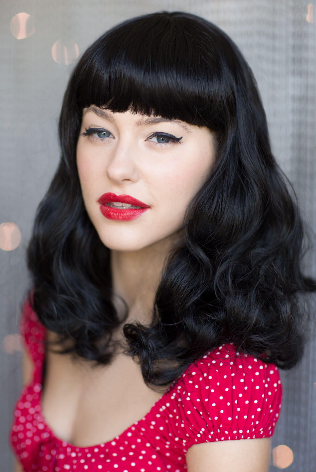 Woman with black hair and red lipstick wearing a red polka dot dress against a neutral background. Black pinup style wig, curled with short fringe, 1950s style: Bettie 1B