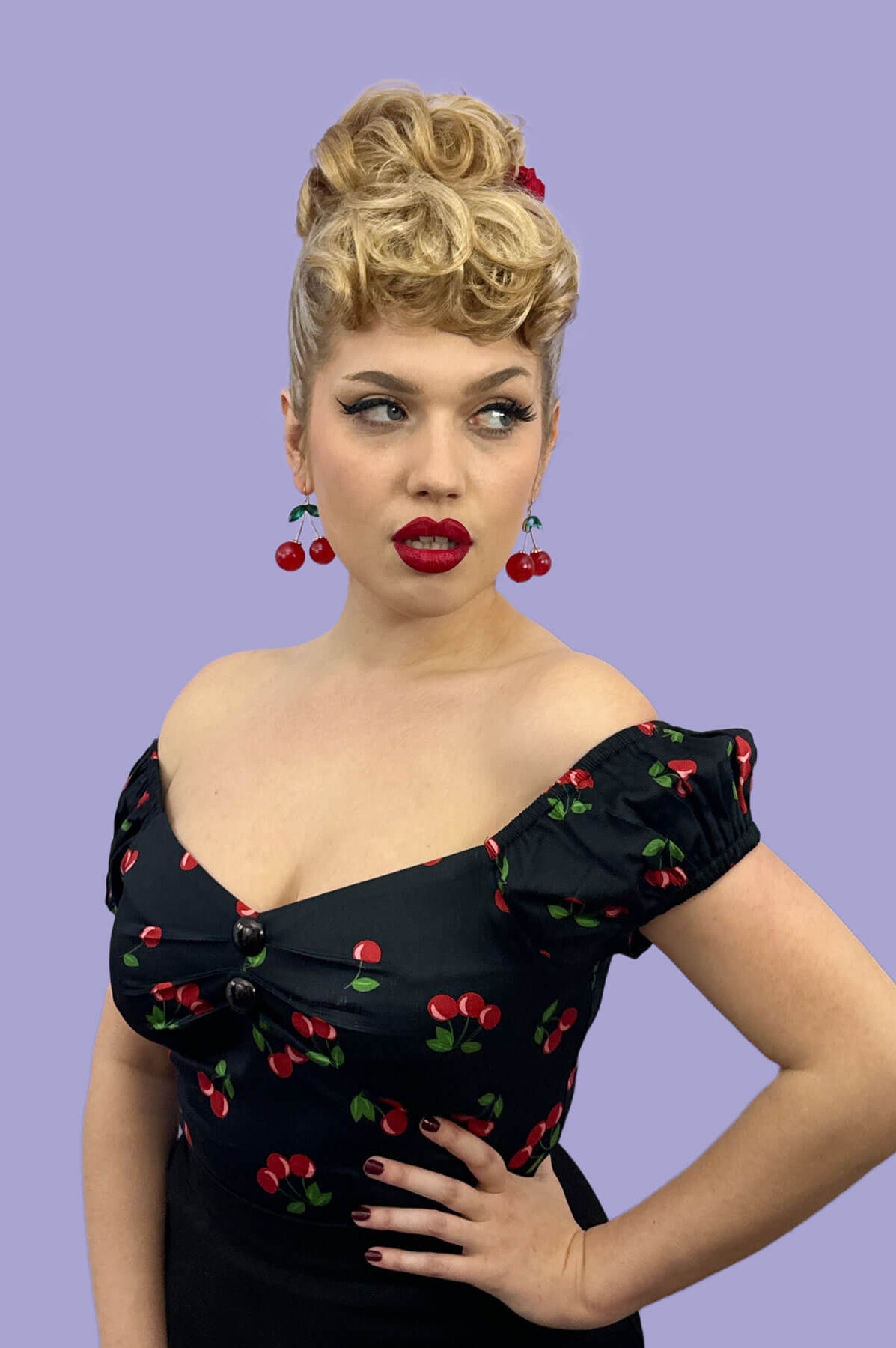 Woman wearing a black dress with red cherry patterns against a purple background. Curly bun hairpiece, rockabilly style: Toni blonde 613T24