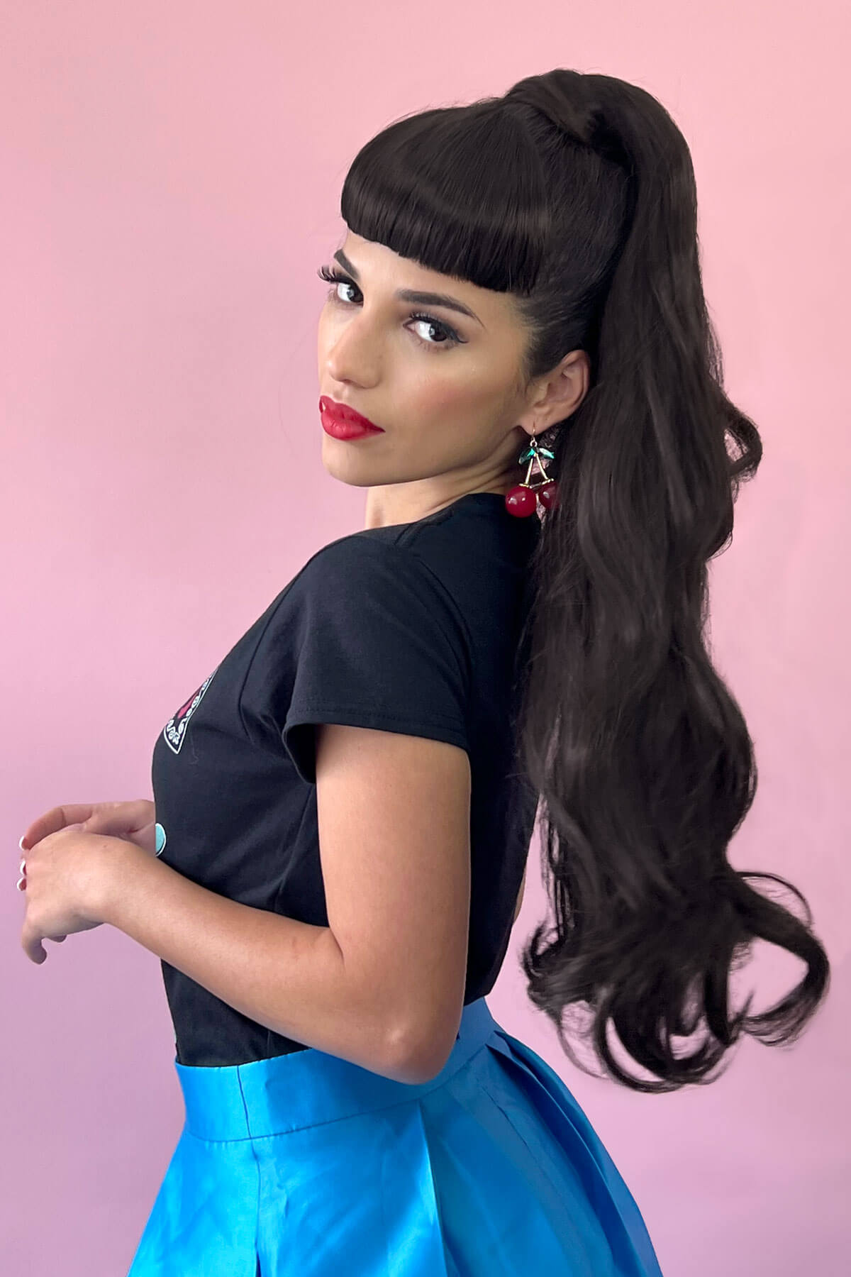 Woman with long dark hair styled in a high ponytail, wearing a black top and blue skirt against a pink background. Sophie ponytail in dark brown