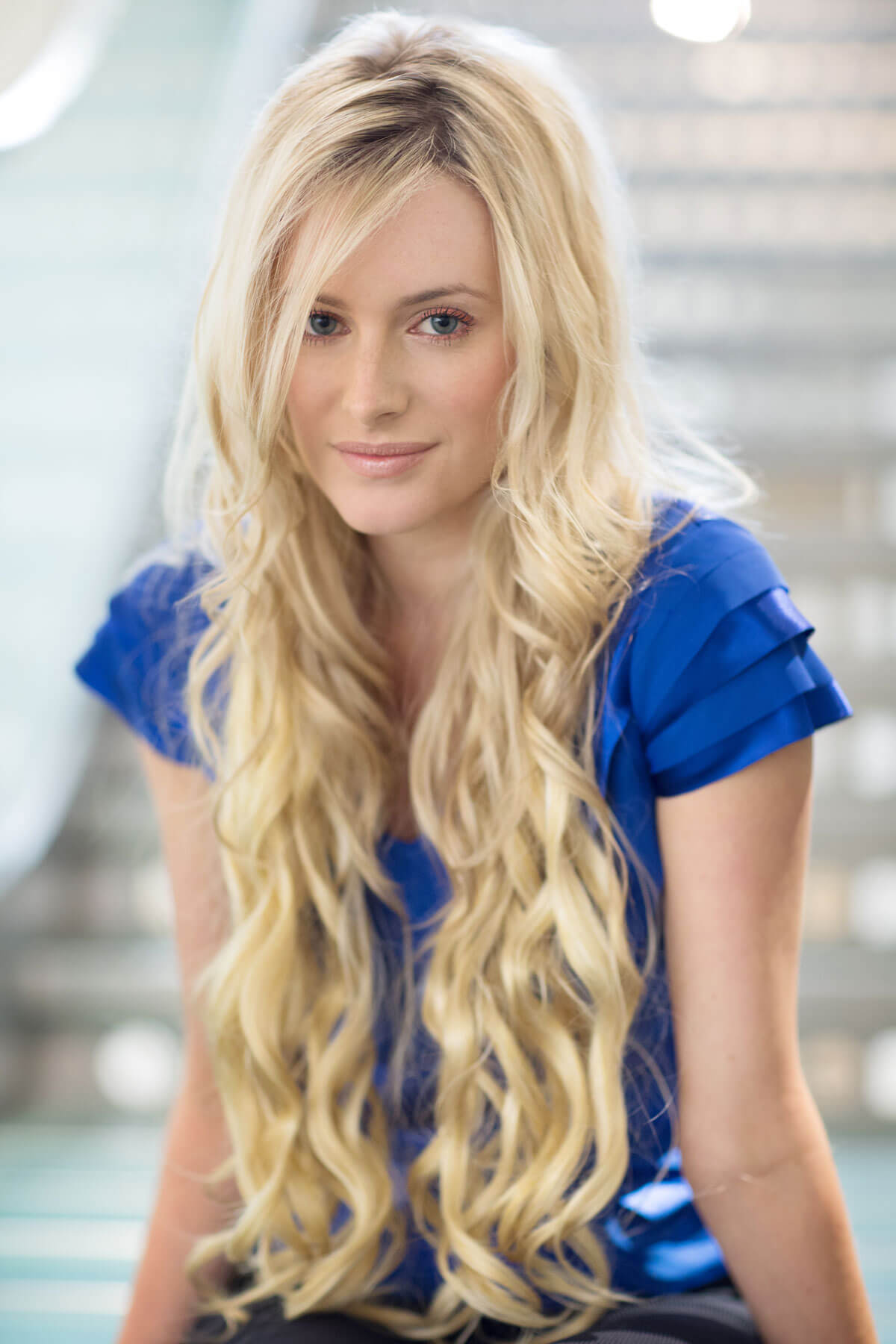 Woman with long blonde hair wearing a blue top in an indoor setting. Extension hairpiece, long, versatile, Flexihair: Cristina wheatfield blonde 24H613