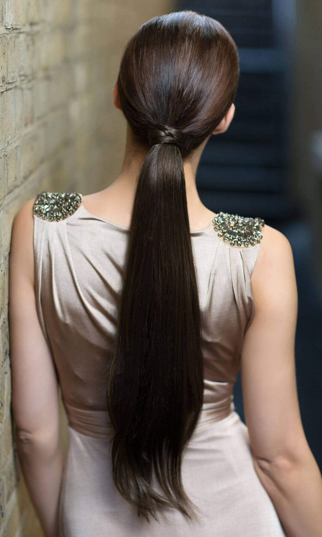 Woman with long dark hair styled in a high ponytail, wearing a light-colored dress with decorative shoulder embellishments. Long sleek ponytail extension with gentle, layered waves: Mali chestnut brown 8
