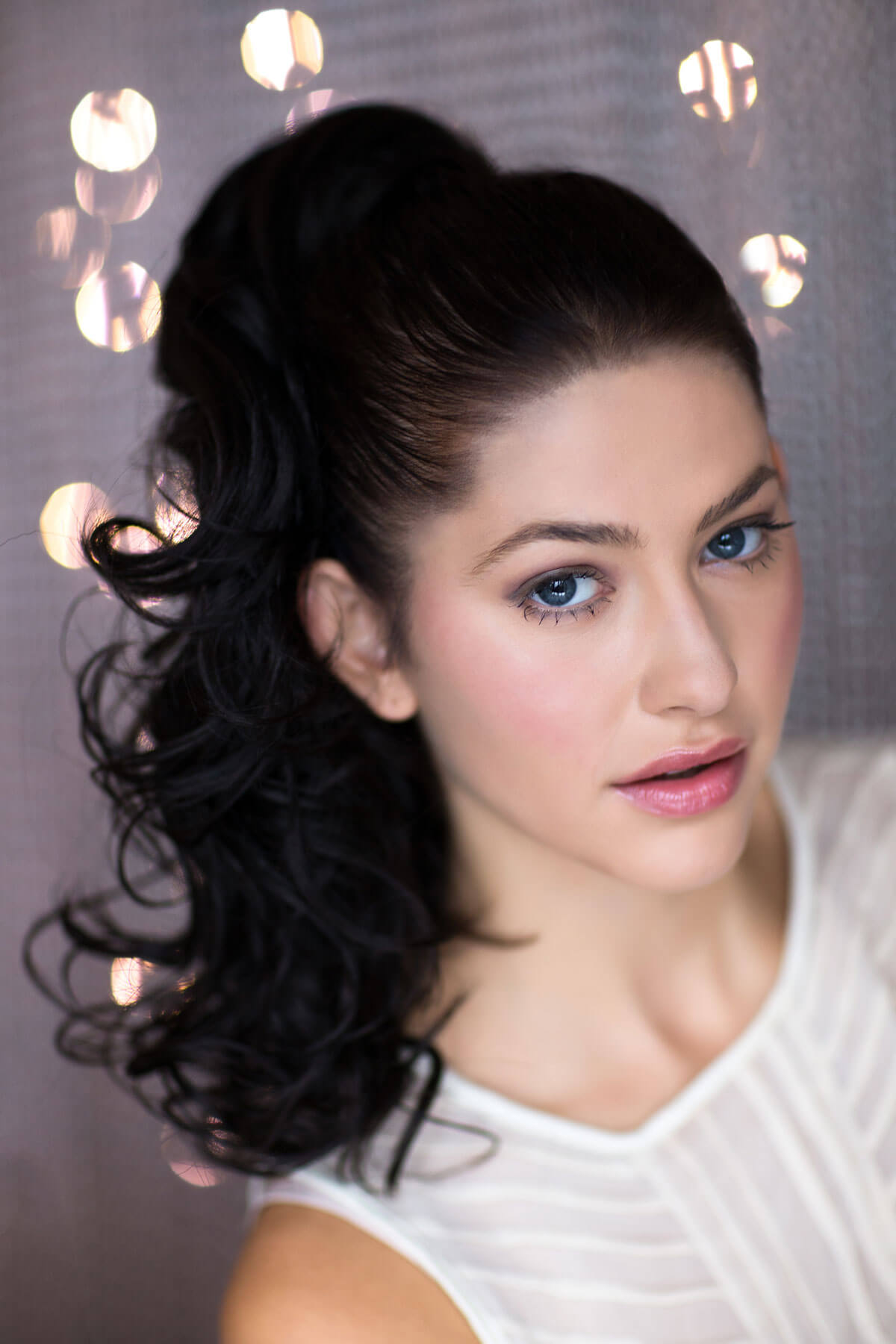 A woman wearing a curly ponytail extension hairpiece with loose, layered curls. She is seen from the shoulders up, with a backdrop featuring bokeh lights. Curly ponytail extension clip-on hairpiece: Tulisa dark brown 4
