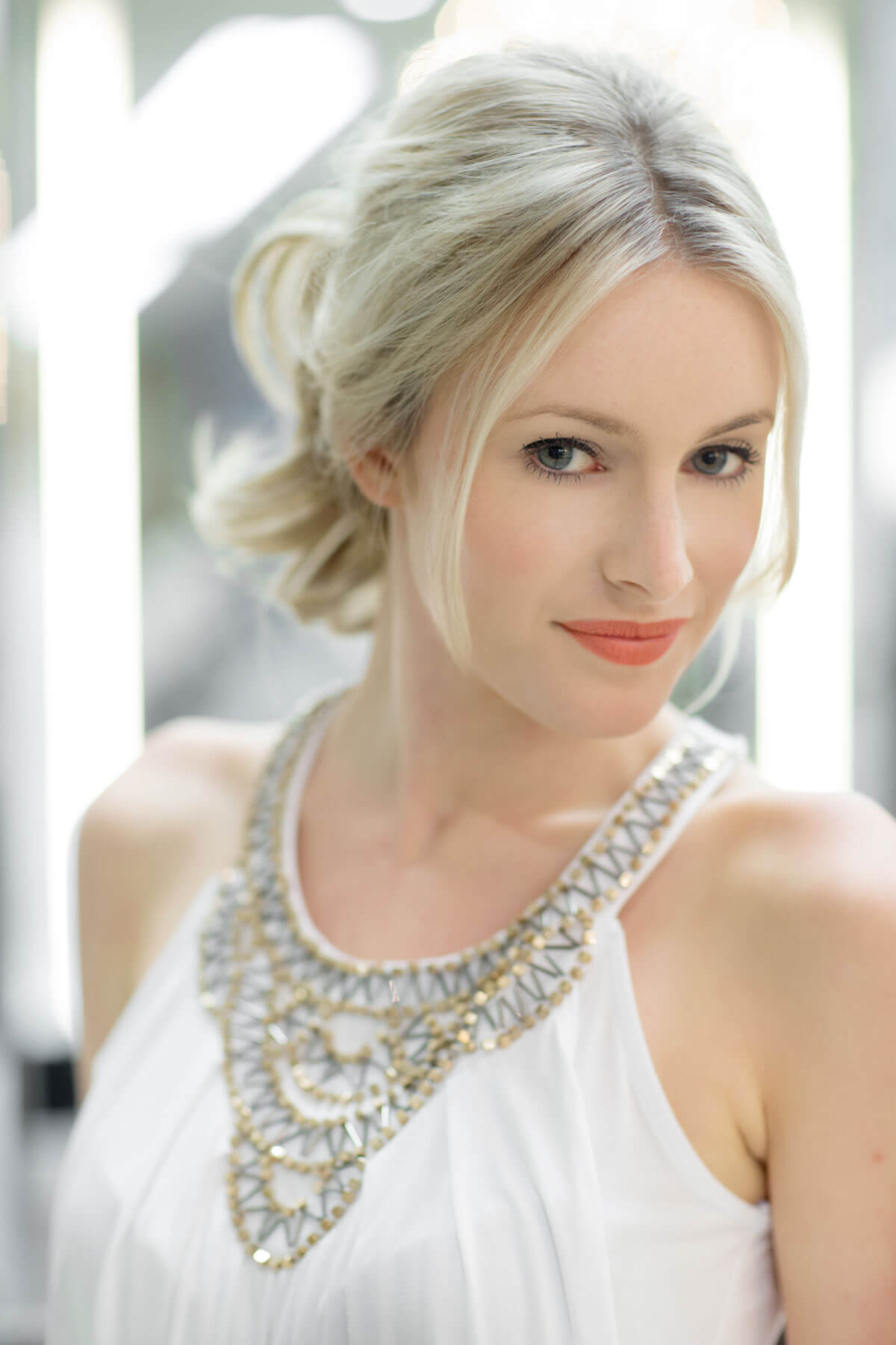 Woman with styled hair and a necklace against a blurred background. Clip in hairpiece, ponytail, short, flexihair: Leela blonde 24H613