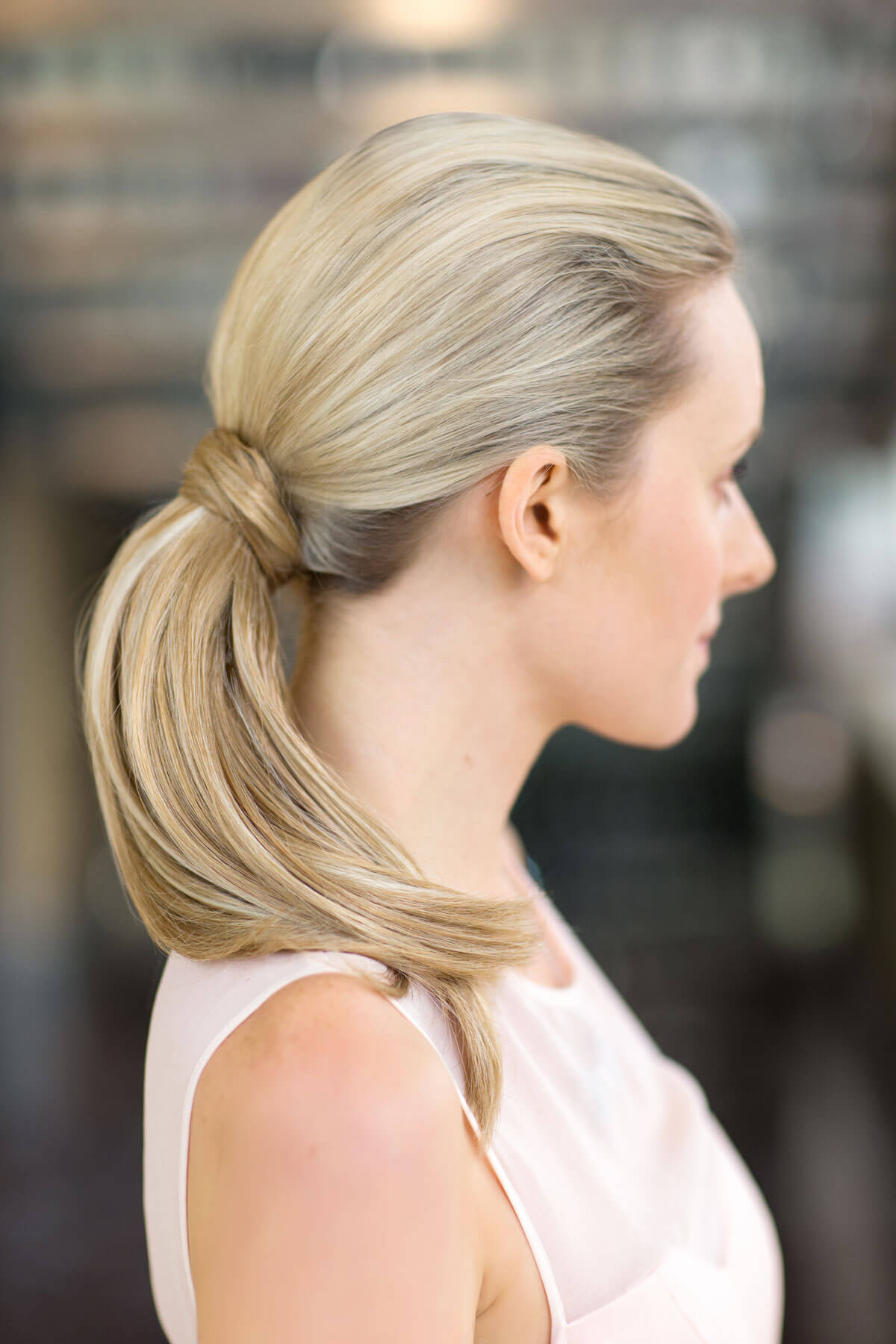 Woman with blonde hair styled in a ponytail against a blurred background. Sleek ponytail extension hairpiece, Flexihair: Jasmin blonde 24H613