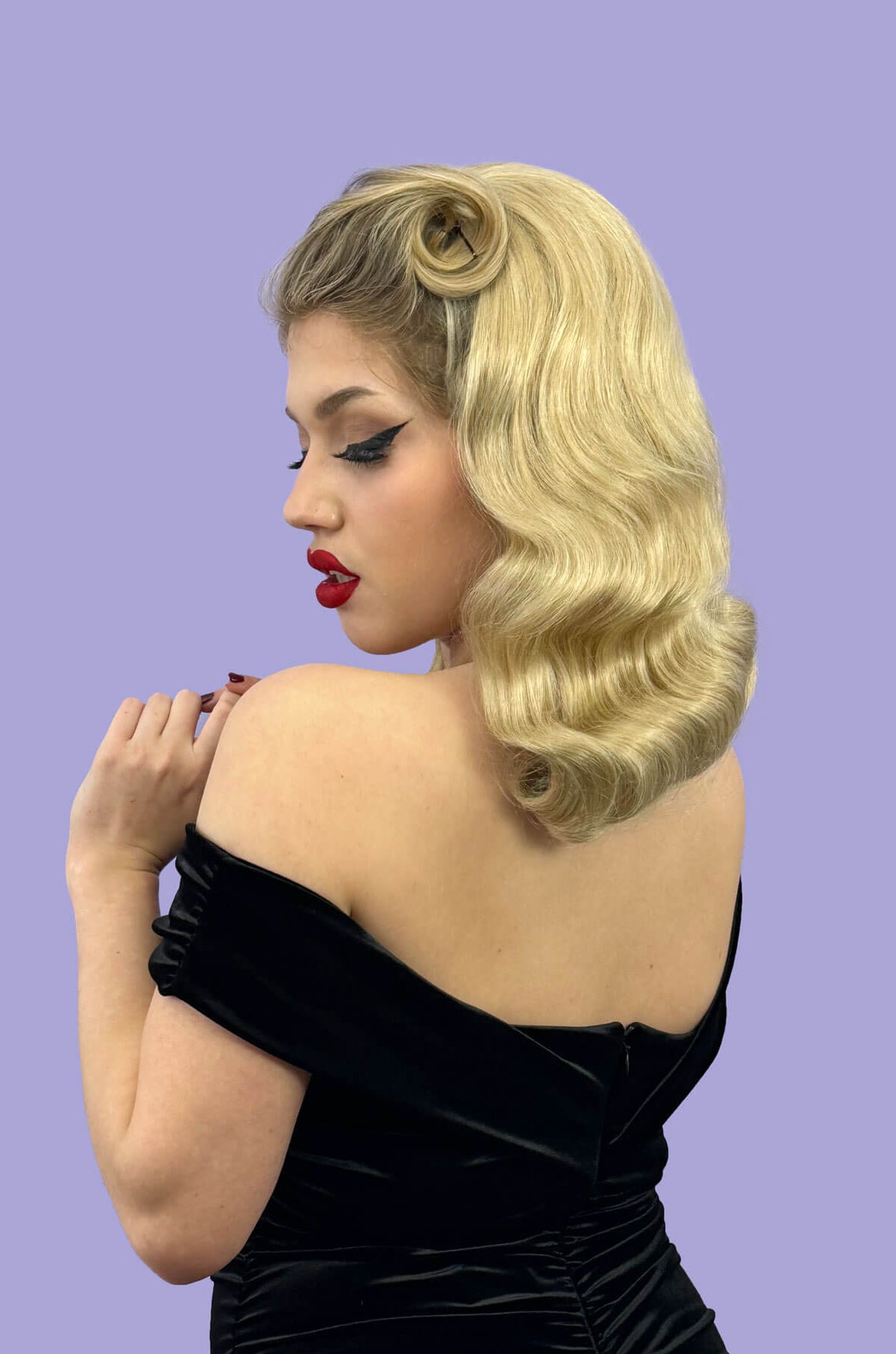 Woman with vintage-style blonde hair and red lipstick wearing a black dress against a purple background. 1950s style half wig hairpiece with deep finger waves: Deborah blonde 613T24