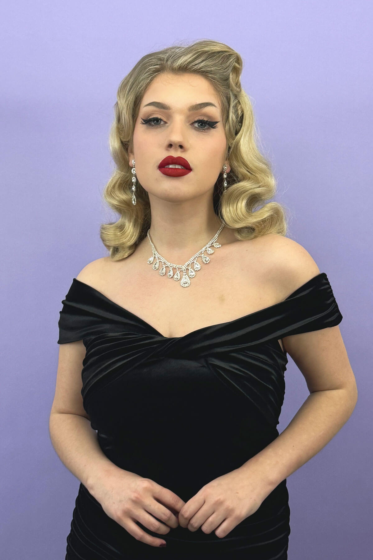 Woman wearing a black off-shoulder dress with a purple background. 1950s style half wig hairpiece with deep finger waves: Deborah blonde 613T24