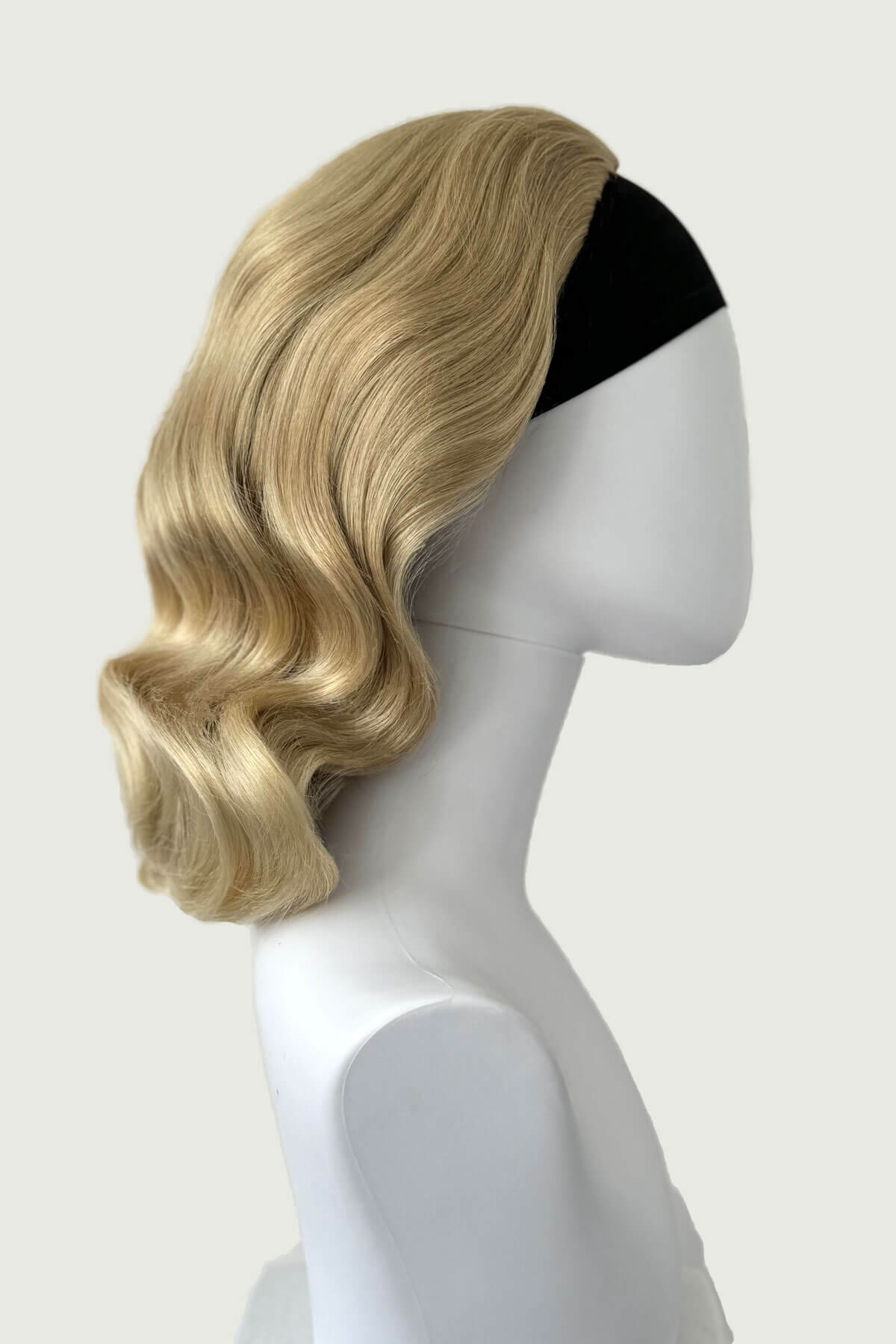 Blonde wig on a mannequin head against a white background, viewed from the side. 1950s style shoulder length half wig hairpiece with beautiful vintage waves: Onyx blonde 613T24