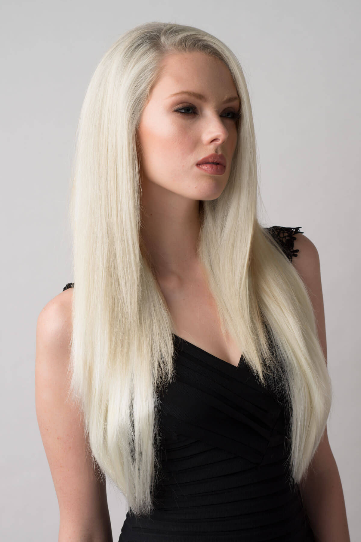 Woman with long blonde hair wearing a black top against a gray background. Straight platinum blonde half wig hairpiece (3/4 wig): Shannon
