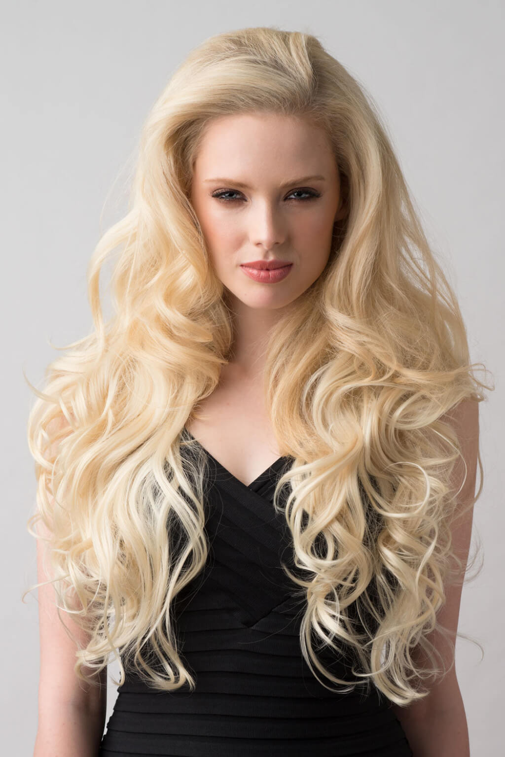 Woman with long, wavy blonde hair wearing a black top against a grey background. Blonde half wig hairpiece (3/4 wig), loose curls: Suki creamy blonde 613