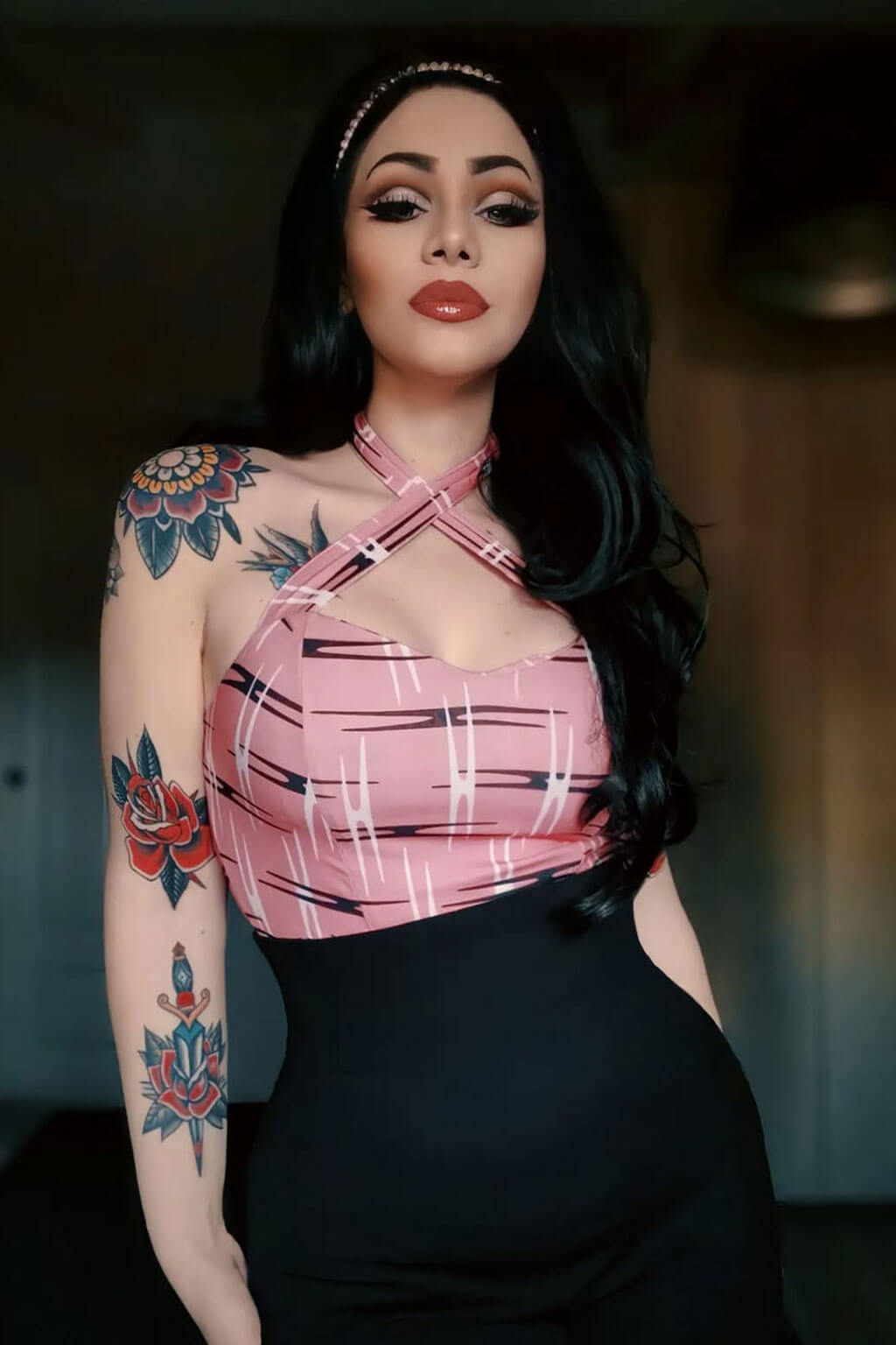 Woman wearing a pink top with black patterns and a black skirt, standing indoors. High crown long, wavy half wig and volume ring: Bardot jet black 1