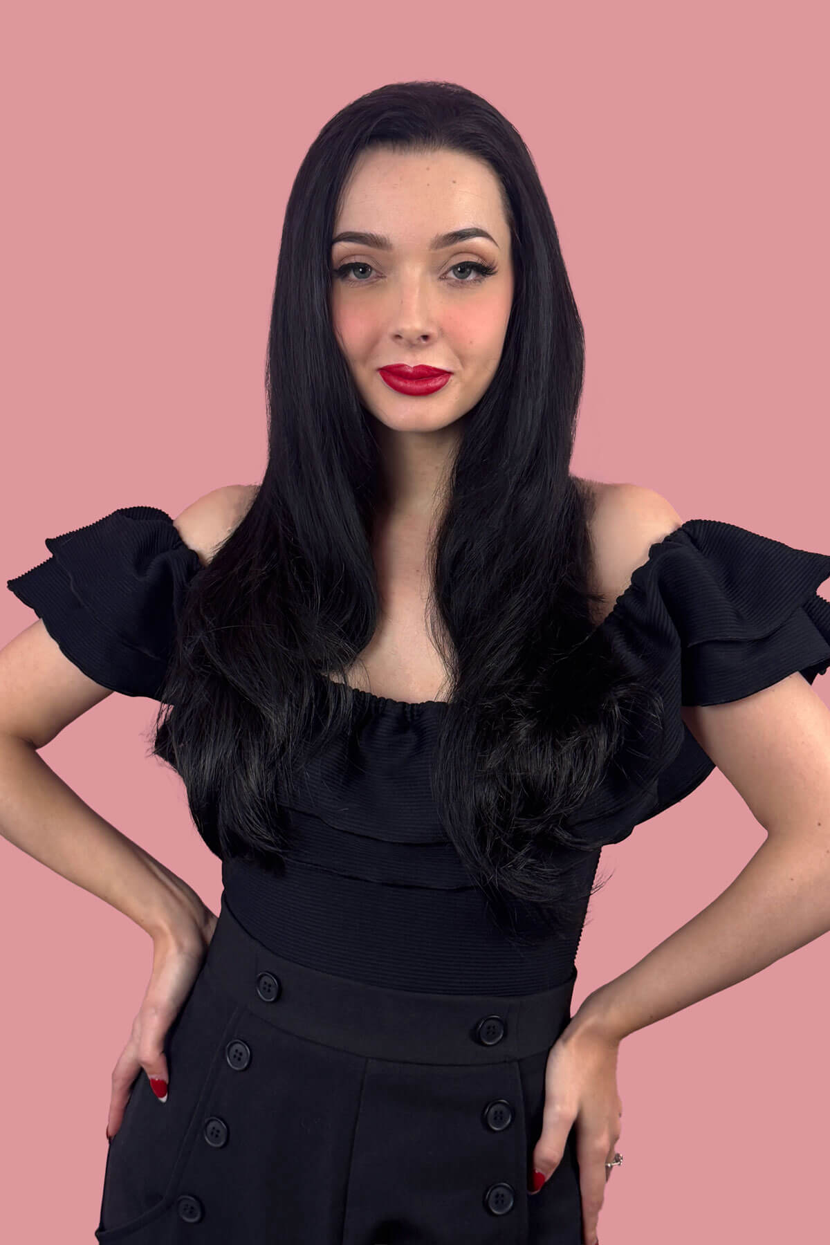 Woman with long black hair wearing a black off-shoulder top with ruffled sleeves against a pink background. Straight black hairpiece (3/4 wig), long and layered: Raven