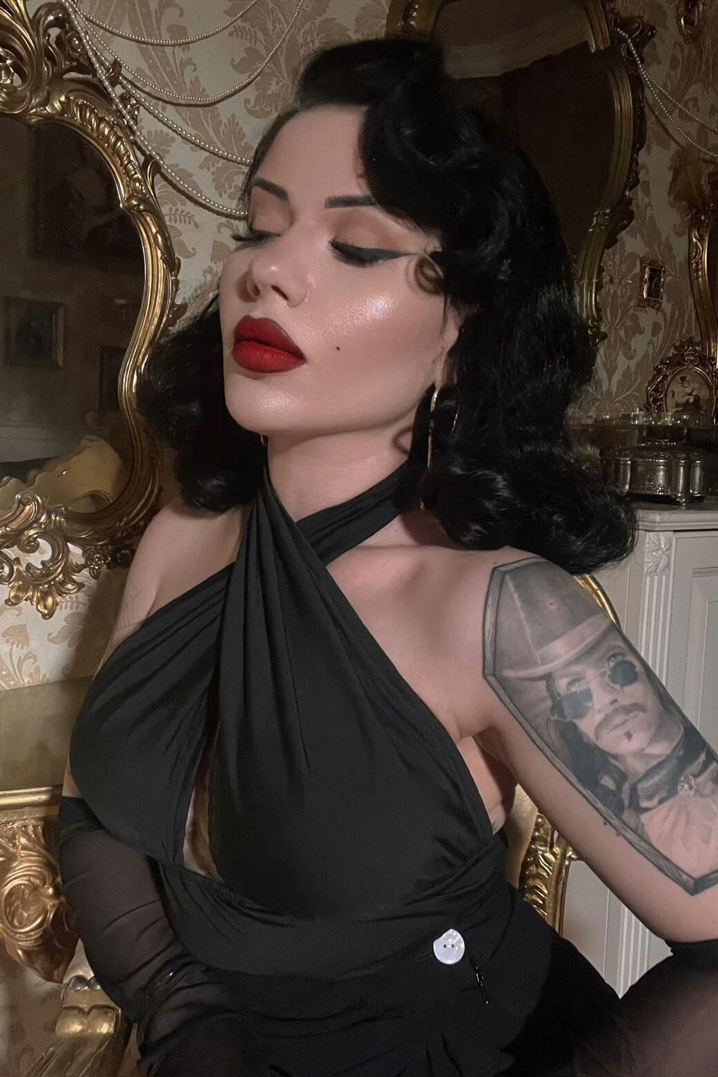 Woman in a black dress with tattoos, sitting in an ornate room. 1940s style half wig hairpiece with beautiful marcel waves: Virginia jet black 1