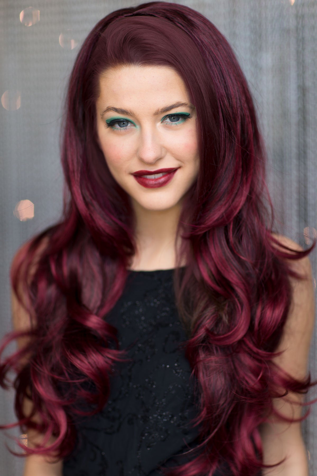 Woman with long, wavy, dark red hair wearing a black top against a neutral background. Black and red half wig hairpiece (3/4 wig), loose curls: Monica
