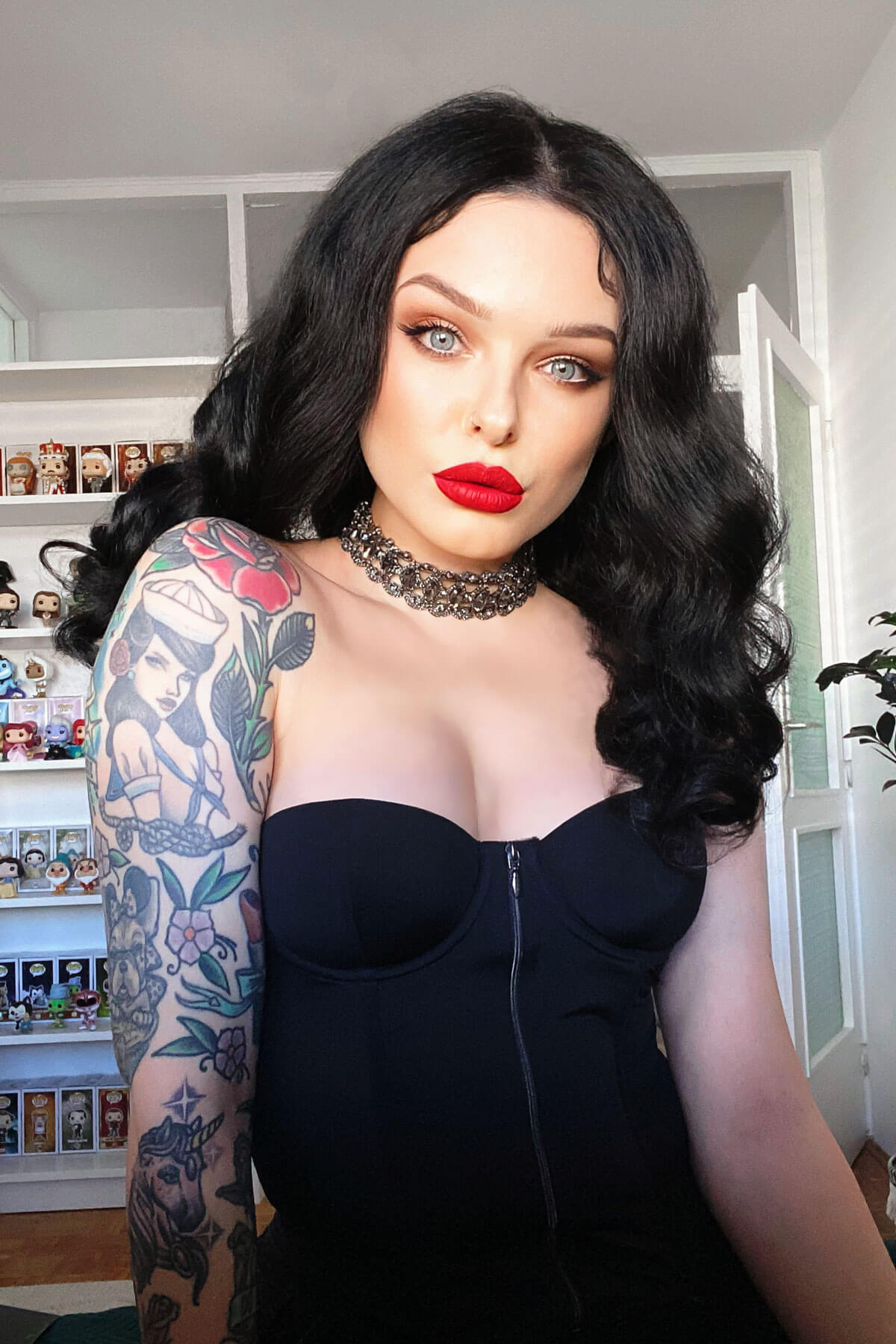 Woman with tattoos and red lipstick wearing a black dress in a room with shelves. Black pinup wig, lace front with long finger waves: Tyra