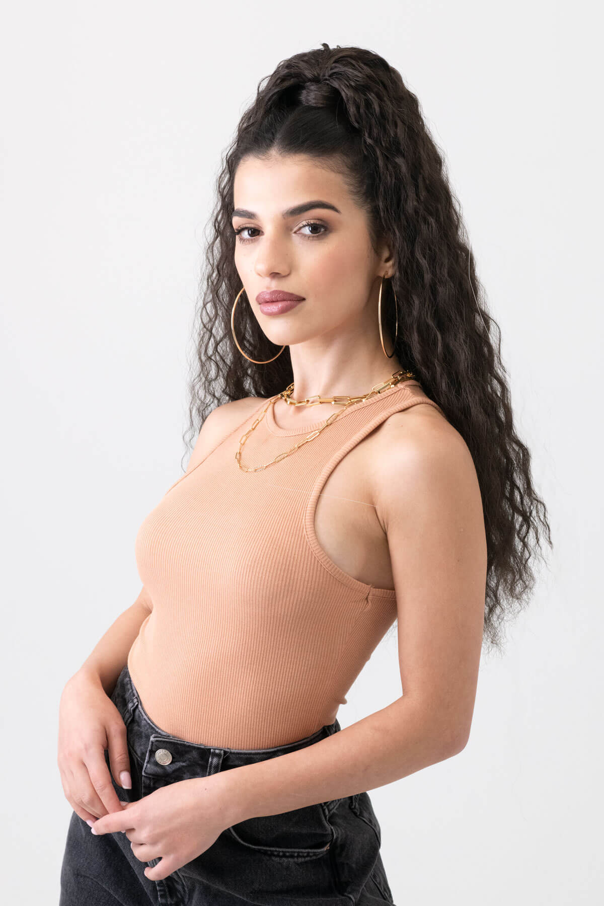Woman wearing a beige tank top and black pants on a white background. Extra long ponytail extension with long loose spirals: Kaia dark brown 4