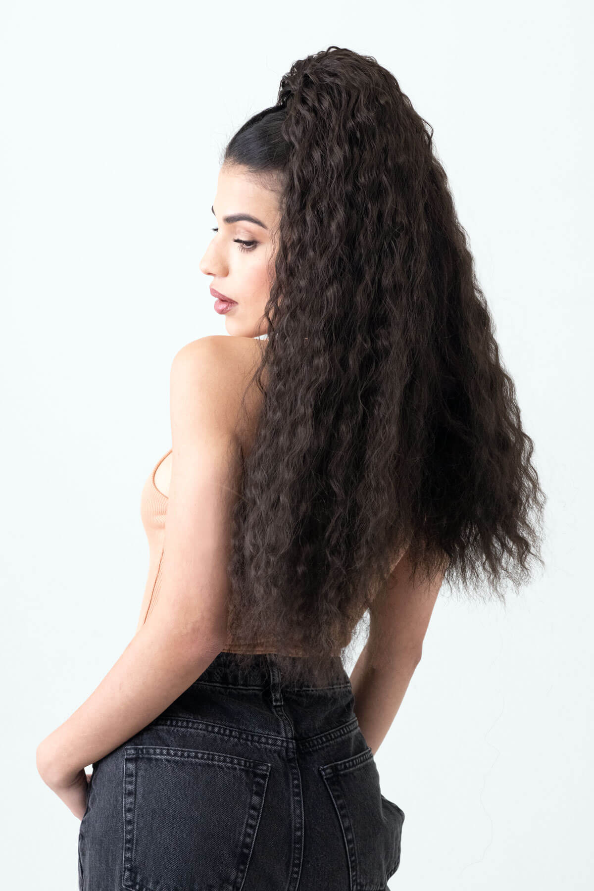 Woman with long, wavy hair styled in a high ponytail on a white background. Extra long ponytail extension with long loose spirals: Kaia dark brown 4