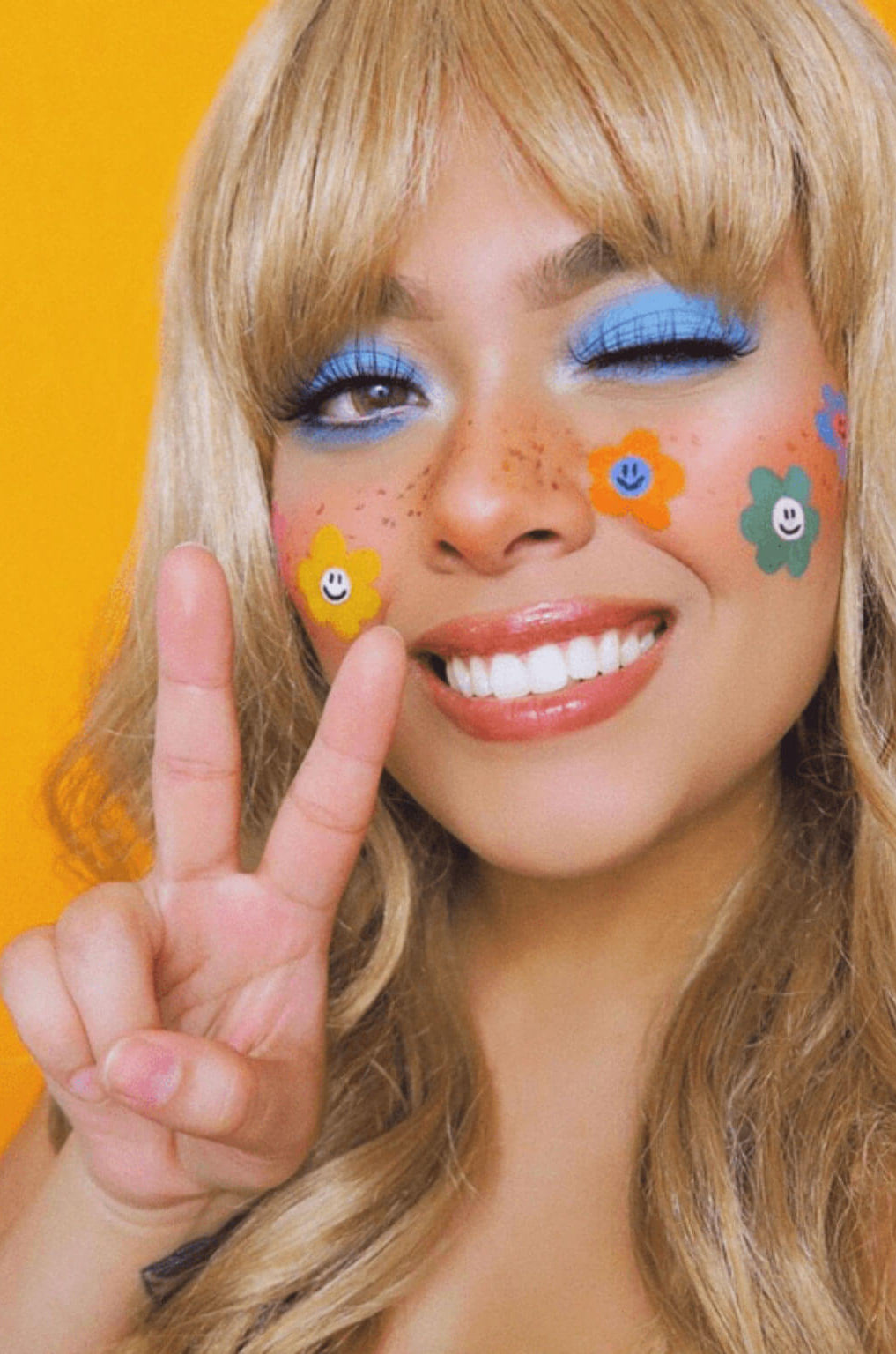 Person with colourful face paint and flowers, making a peace sign against an orange background. Lucky dip wig