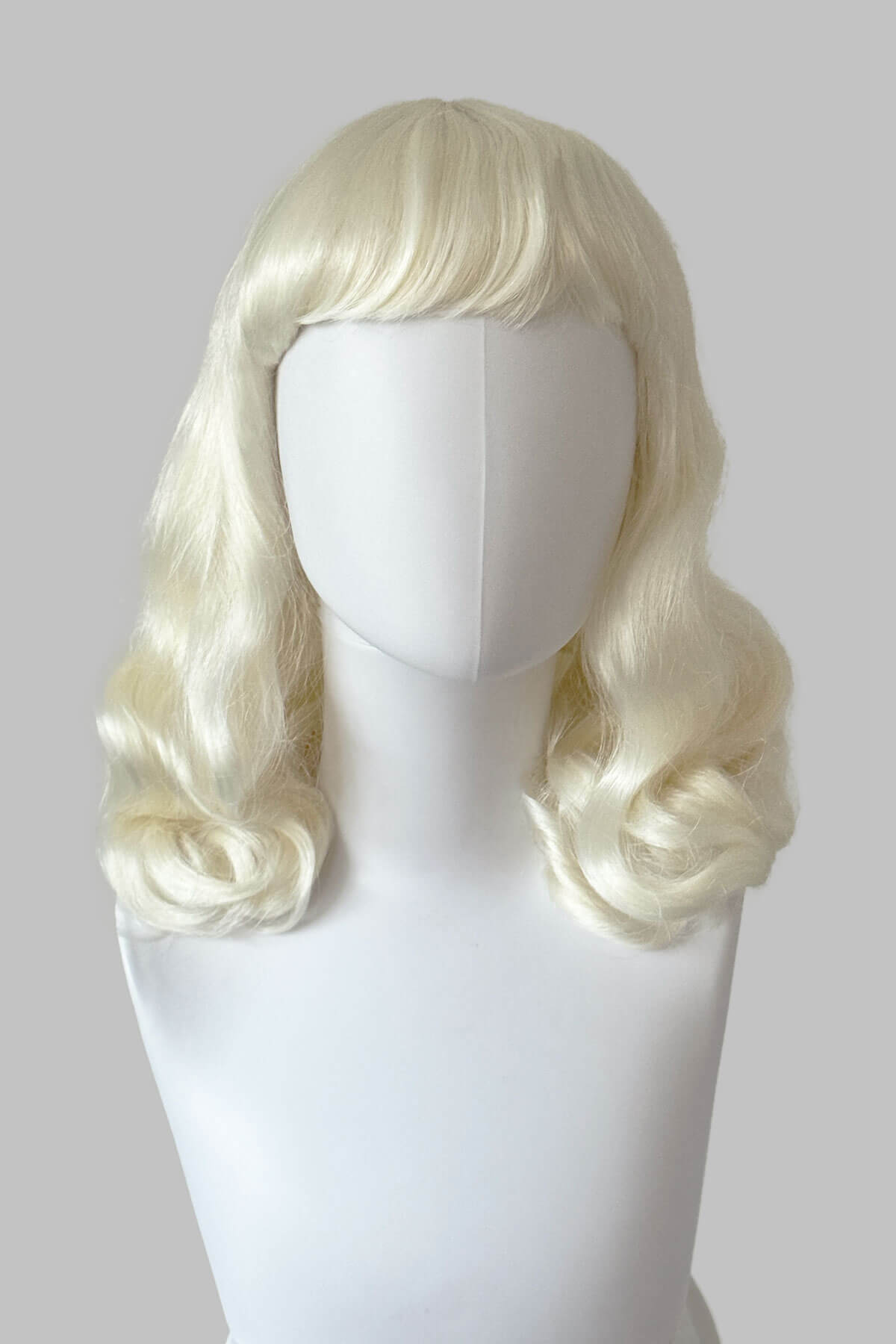 Blonde wig on a mannequin head against a plain background. Platinum blonde pin-up style wig with short fringe and marcel waves: Eira