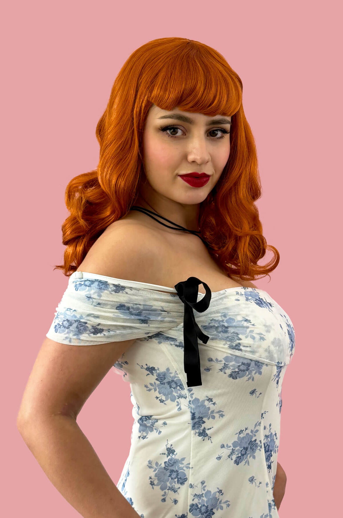 Woman with red hair and a floral off-shoulder dress against a pink background. Ginger pinup style wig, finger waved with short fringe, 1950s style: Sienna