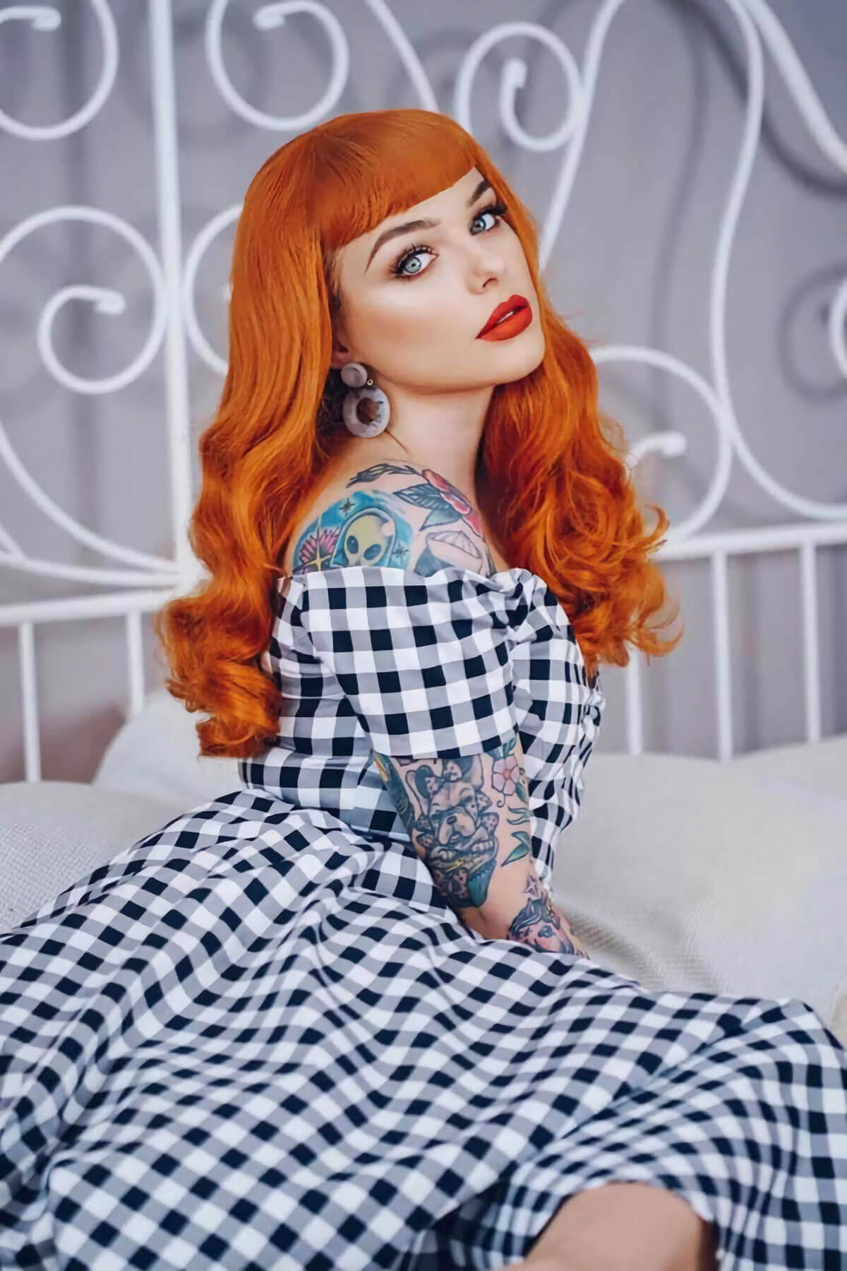 Woman with red hair and tattoos wearing a checkered dress sitting on a bed. Ginger wig, pinup style with short fringe and gentle waves, 1950s style: Vida