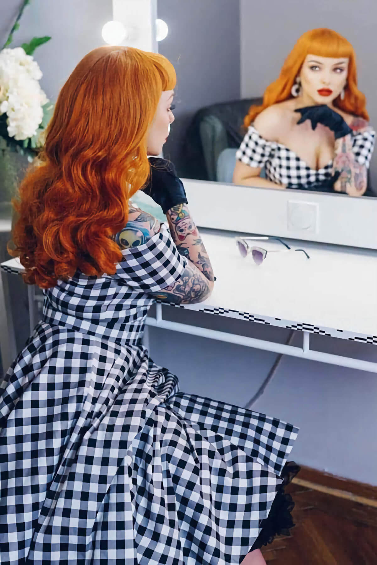 Woman with red hair and tattoos wearing a black and white checkered dress, sitting in front of a mirror. Ginger wig, pinup style with short fringe and gentle waves, 1950s style: Vida