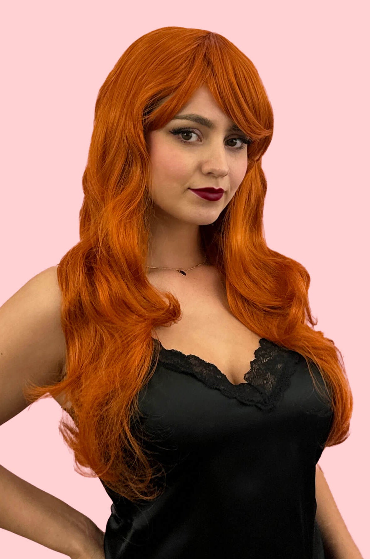 Woman with long orange wig wearing a black top on a pink background. Long ginger wig with loose waves and curls: Ginger