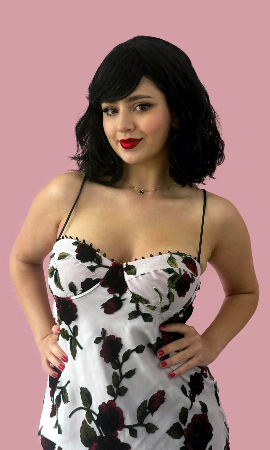 Woman wearing a floral dress against a pink background. Black long wavy bob (lob) wig with swept fringe: Eleena