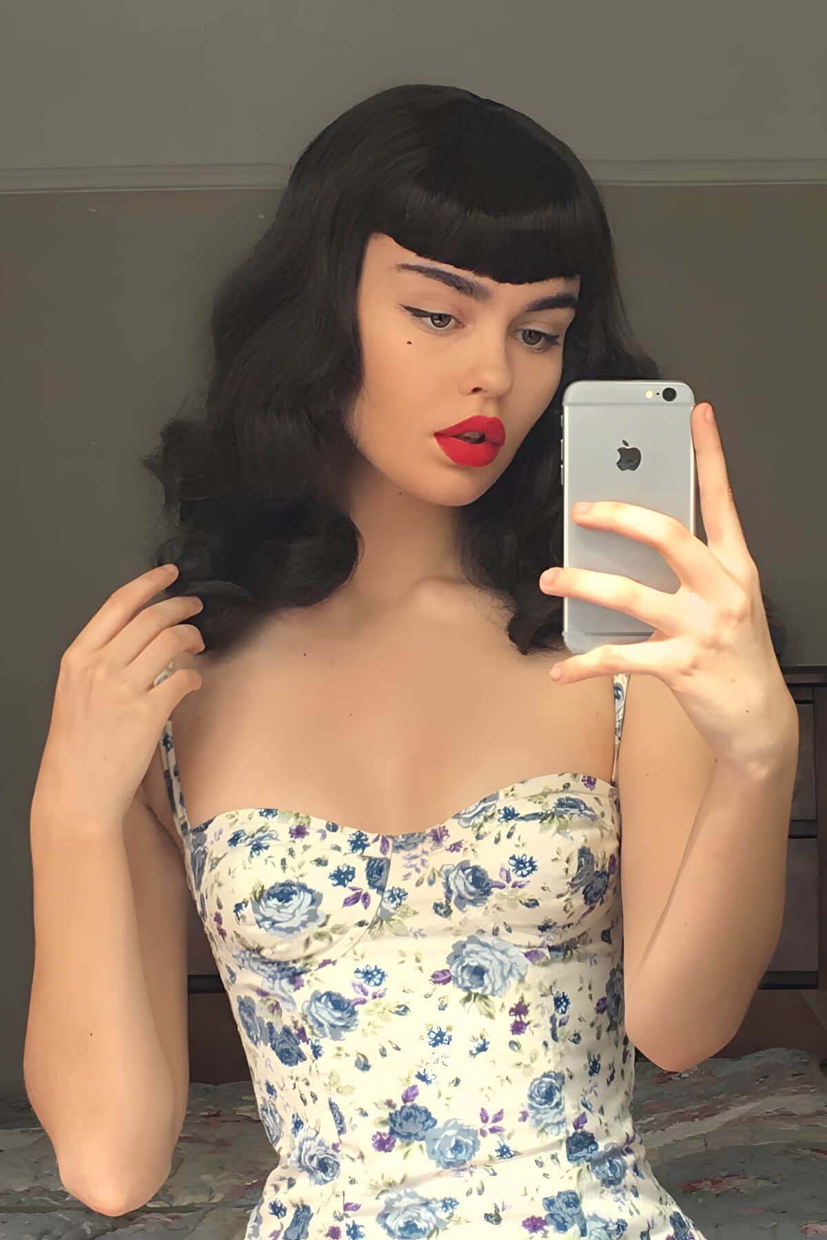 Woman with black hair and red lipstick taking a mirror selfie, wearing a floral dress. Black pinup style wig, curled with short fringe, 1950s style: Bettie