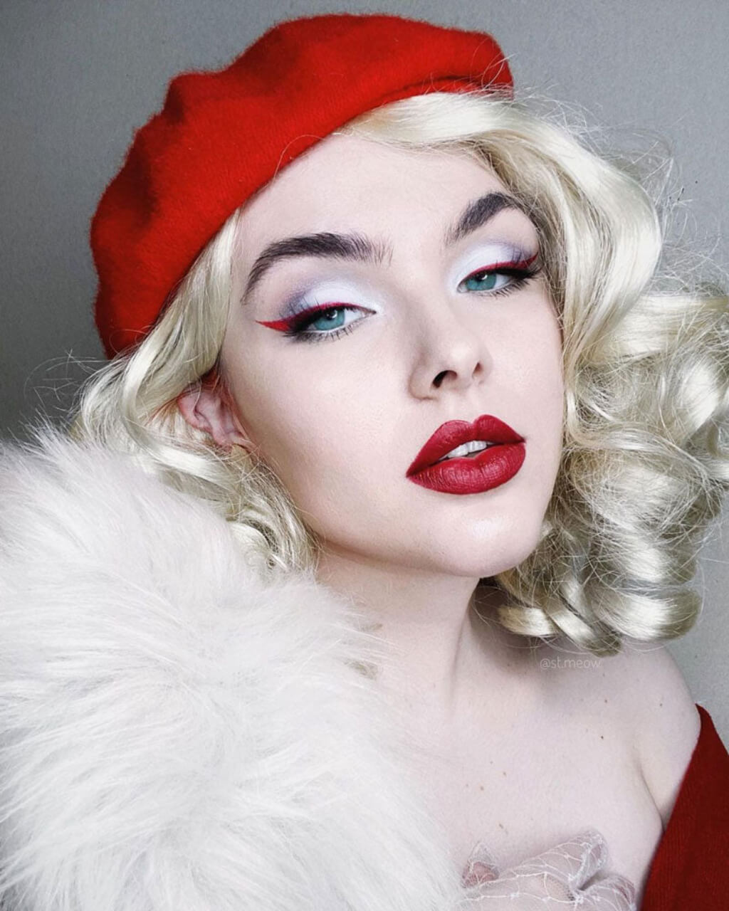 Woman wearing a red beret and white furry collar against a grey background. 40s blonde pinup style wig with marcel waves: Lauren
