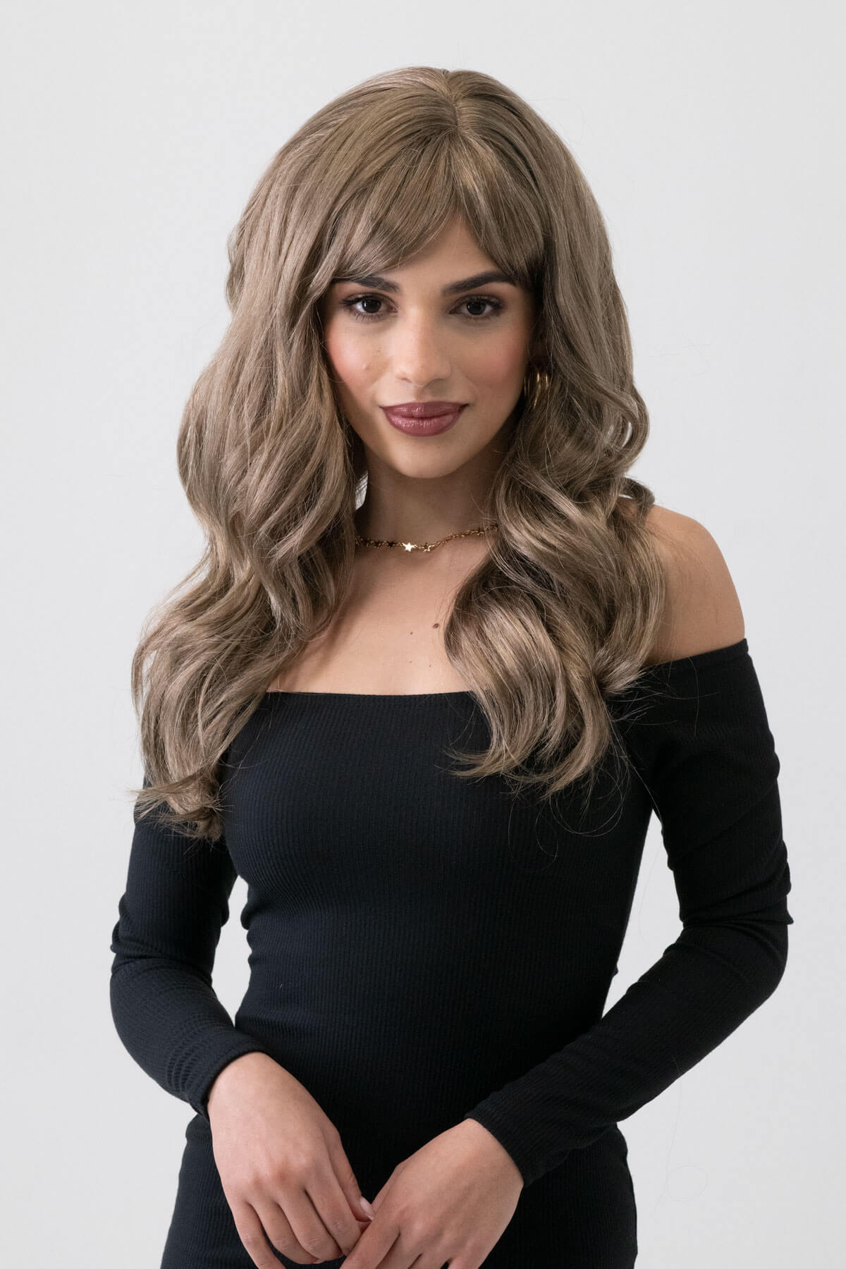 Woman with styled hair wearing a black off-shoulder top on a white background. Light brown wavy wig with ash blonde highlights: Cata