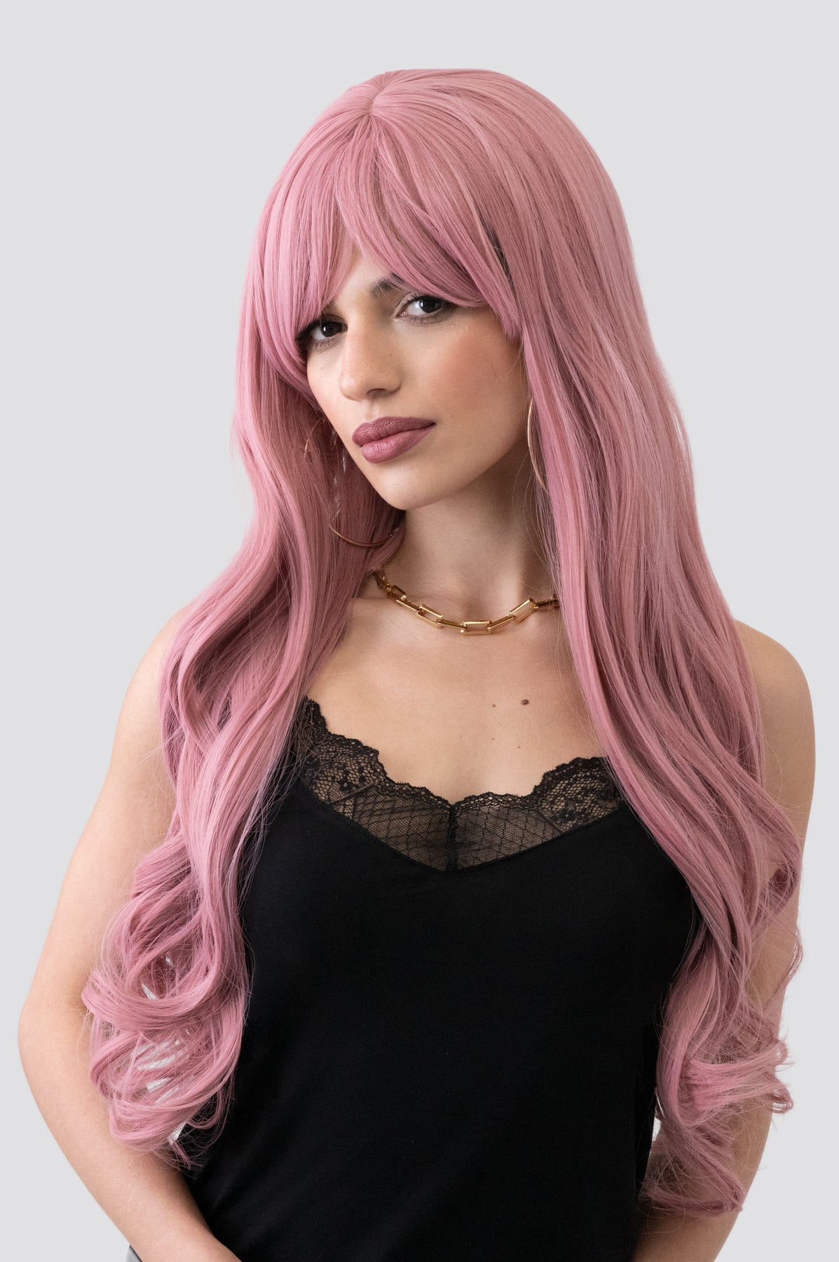 Woman with long pink wig wearing a black top on a light gray background. Long wavy pink wig with sweeping fringe: Millicent