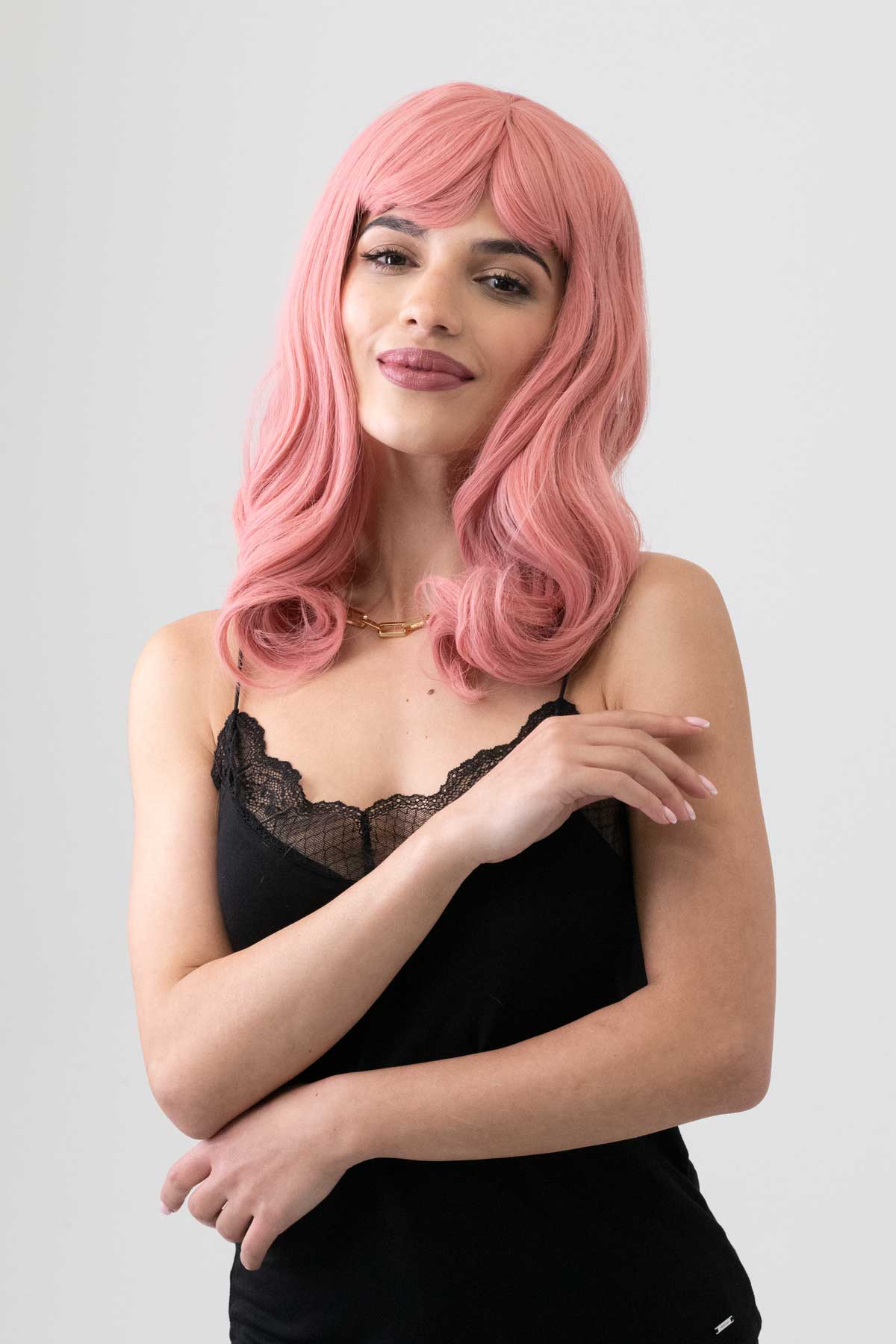 Woman with pink wig wearing a black top on a light gray background. Dusty Pink 50s vintage style wig with short fringe: Paloma