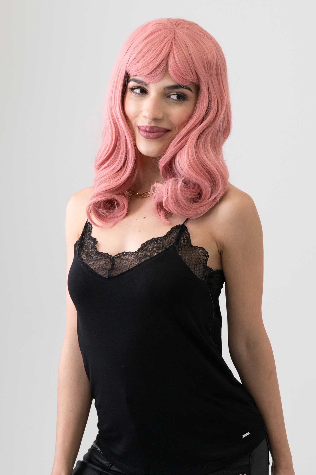 Woman with pink wig wearing a black top on a gray background. Dusty Pink 50s vintage style wig with short fringe: Paloma