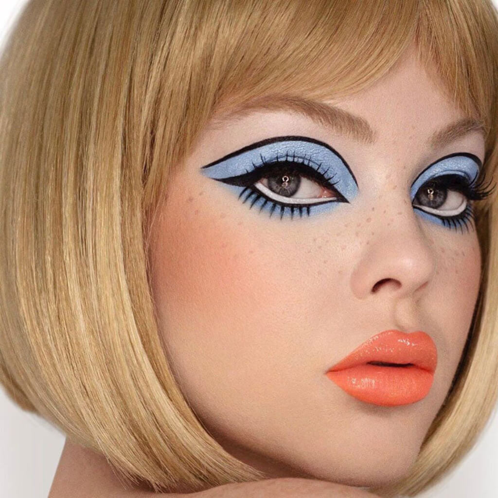 Close-up of a person with blonde hair and dramatic makeup, including blue eyeshadow and orange lipstick. Blunt blonde bob wig with fringe: Gracie 24H613