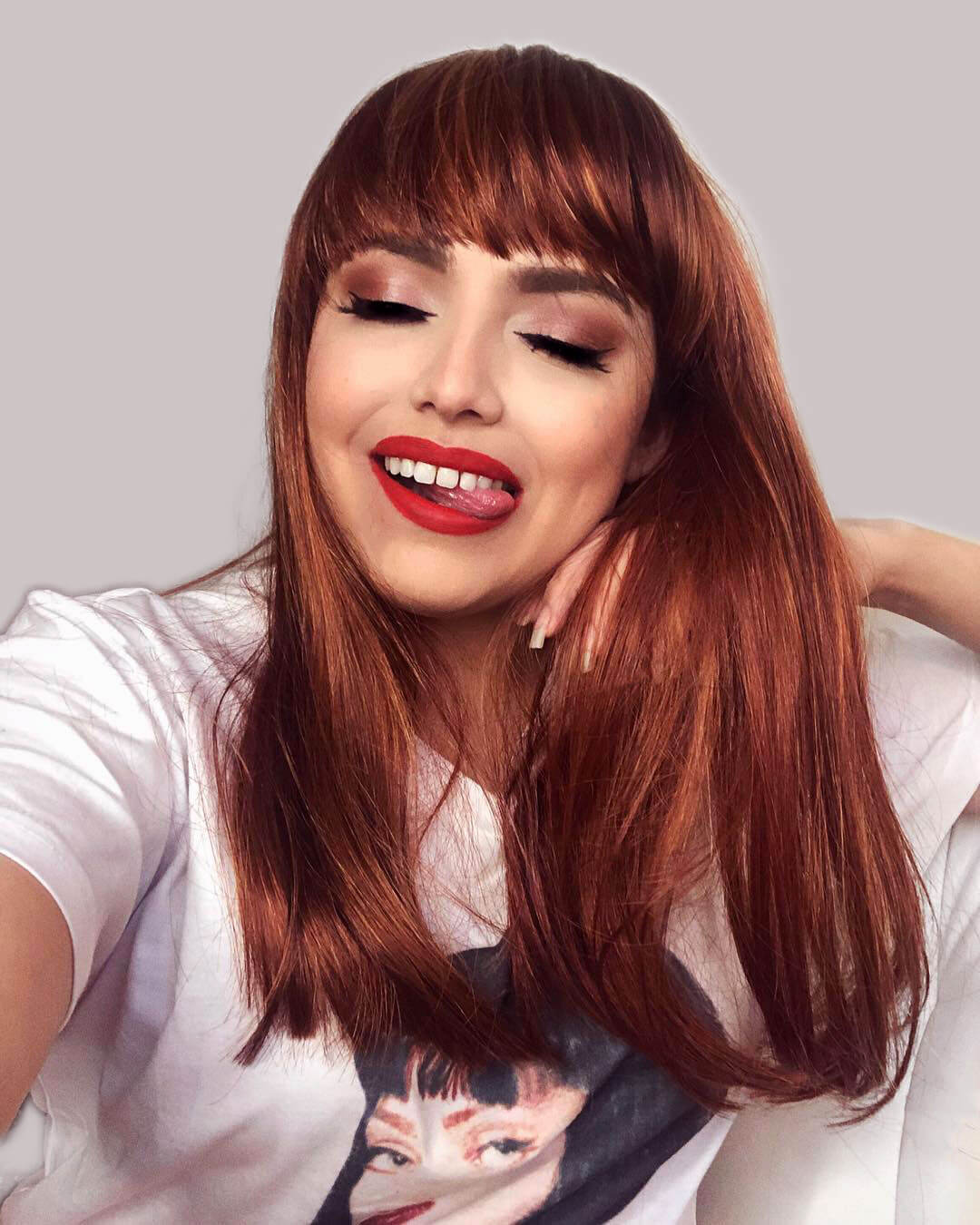 Woman with long red hair and red lipstick smiling against a light gray background. Red/Auburn wig with blonde highlights and straight fringe: Abi