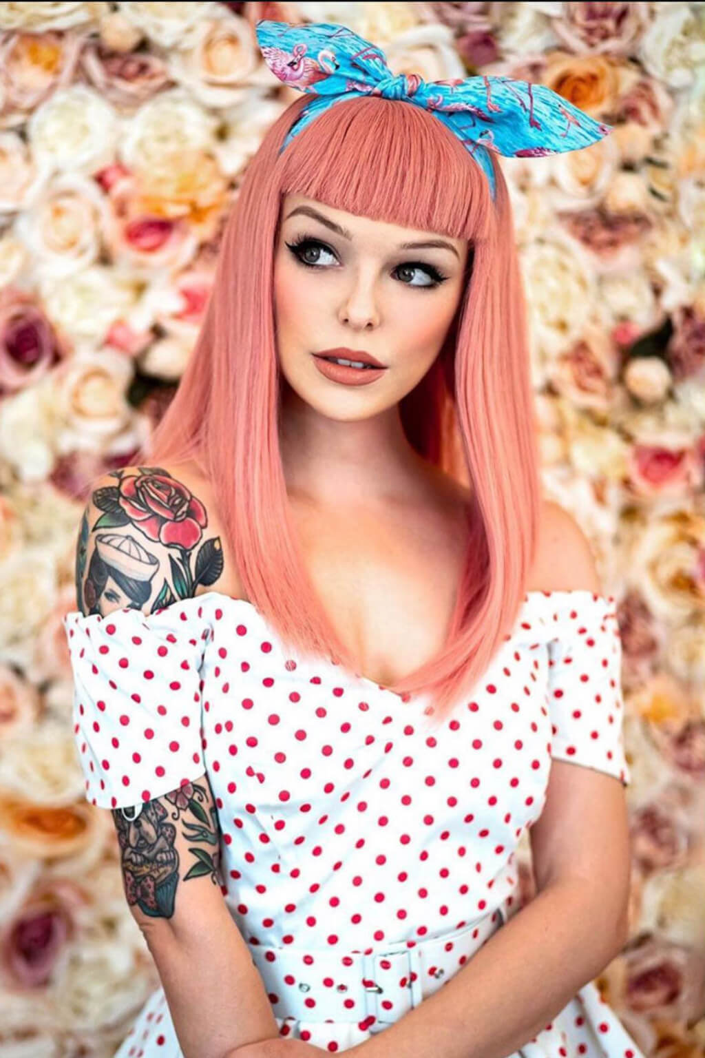 Woman with pink hair and tattoos wearing a polka dot dress in front of a floral background. Long Pink Wig With Short, Straight Fringe: Stephanie