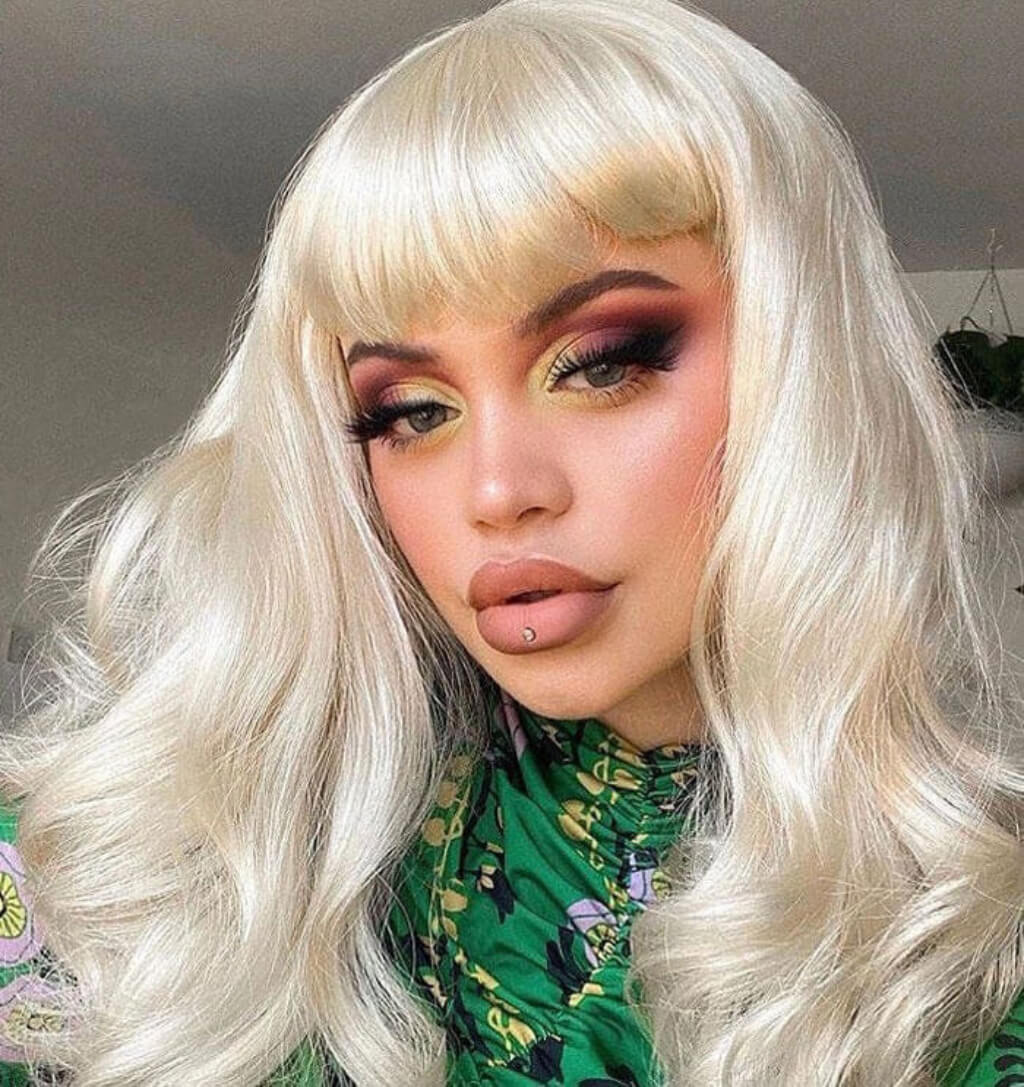 Person with long blonde hair and green floral top. Blonde pinup wig, curled with short fringe, 1950s style: Cora creamy blonde 613