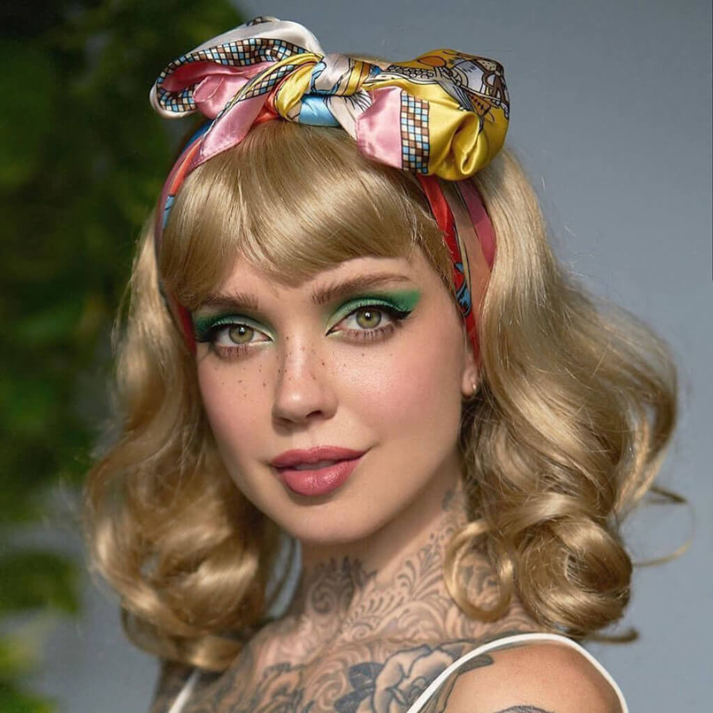 A close-up portrait of a woman with blonde wavy hair wearing a colourful patterned silk headscarf tied in a bow, styled in a retro pin-up look with green eyeshadow and tattoos visible on her neck and shoulder. Blonde pinup wig, curled with short fringe, 1950s style: Cora blonde 24H613