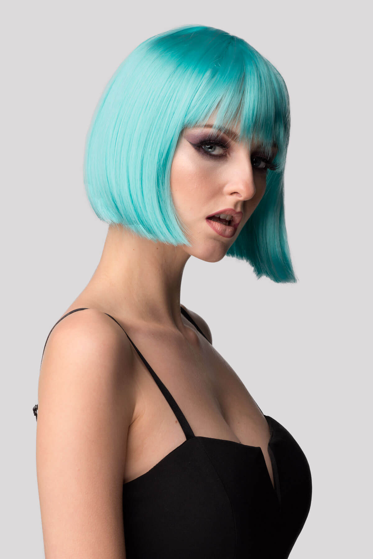 Woman with turquoise hair wearing a black top against a gray background. Turquoise bob wig with full fringe: Iris
