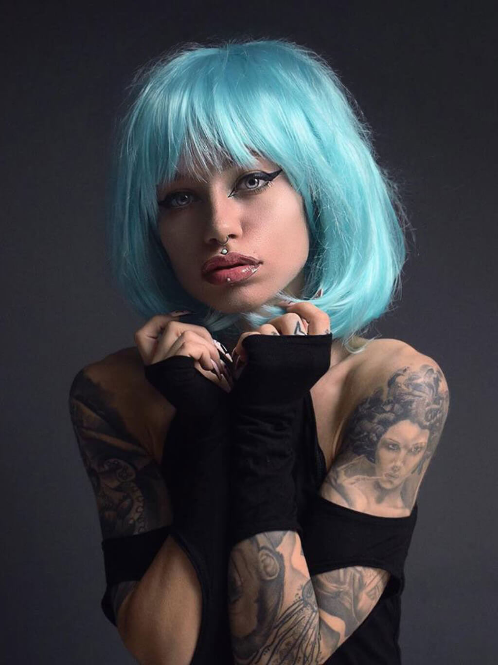 Person with blue hair and tattoos wearing a black outfit against a dark background. Turquoise bob wig with full fringe: Iris