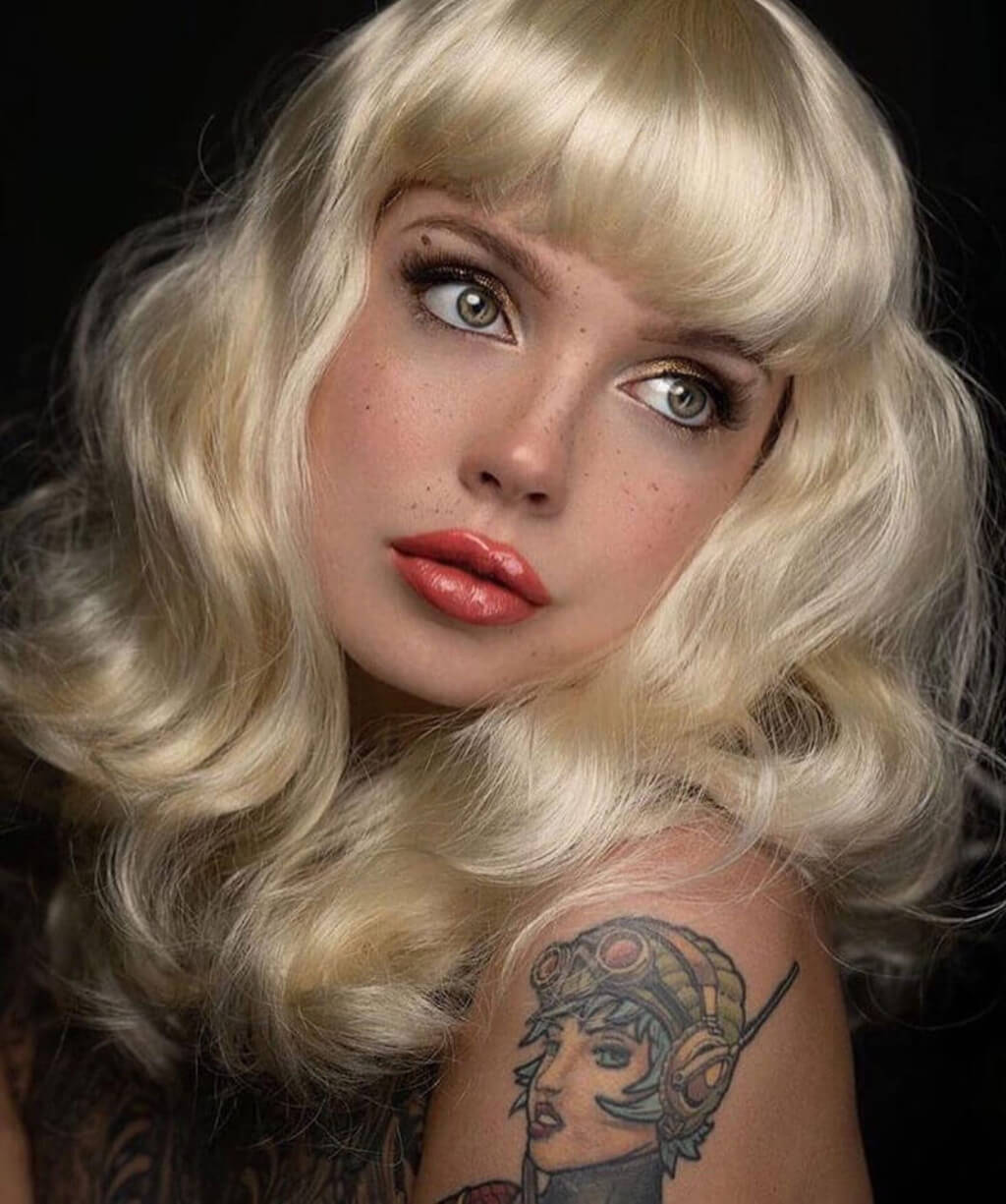 Woman with blonde wig and tattoos on arm against a dark background. Blonde pinup wig, curled with short fringe, 1950s style: Cora creamy blonde 613