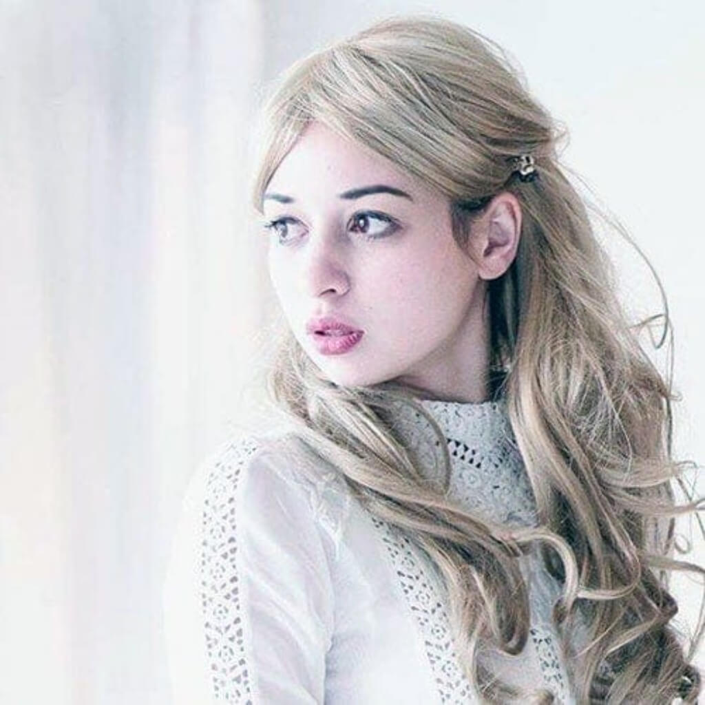 Woman with long blonde hair styled in a side ponytail against a white background. Light blonde wig with big loose curls, extra long: Eva