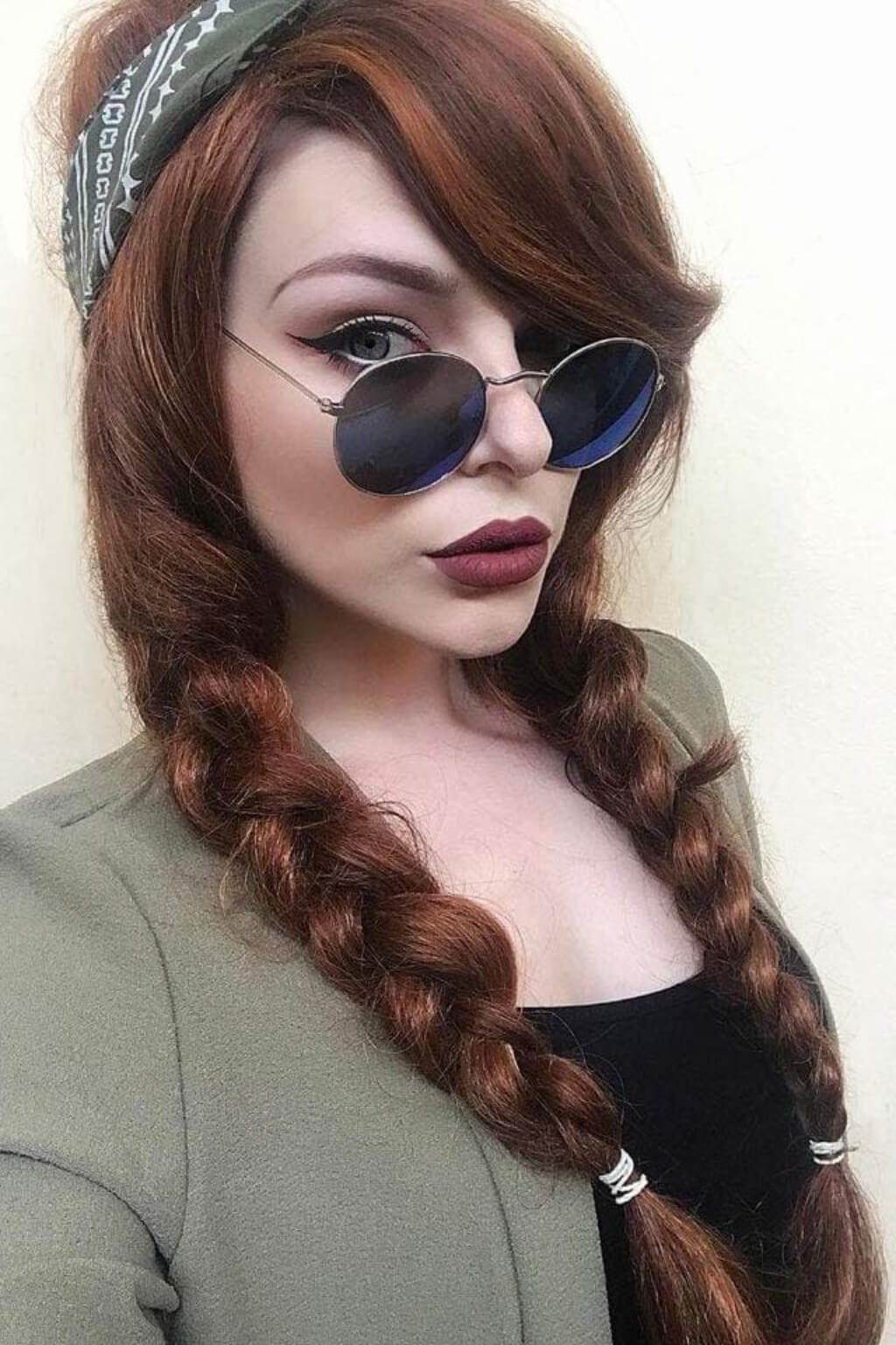 A close-up portrait of a woman with long auburn hair styled in two braids, wearing round dark sunglasses, a patterned headband, and an olive green blazer over a black top. Red & auburn wig with blonde highlights, long: Freya