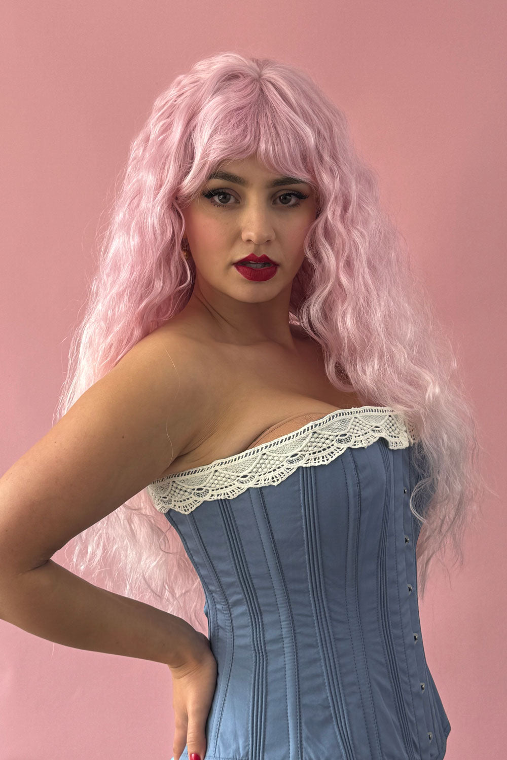 A person wearing a long, crimped pink wig with ombre coloring that gradually blends from dark pink to peach at the tips. The wig wearer is posed against a pink background. Crimped pink wig, long, in two colour pink ombre: Peony