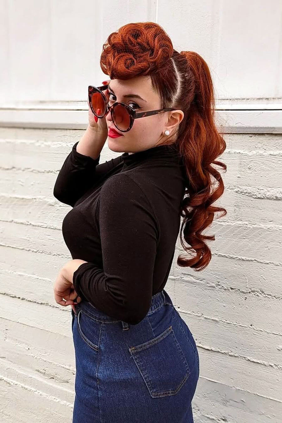 Woman with styled red hair wearing sunglasses and a black top against a light wooden panel background. Hair poodle clip-in hairpiece bun vintage style 1950s: Lucille copper red 130