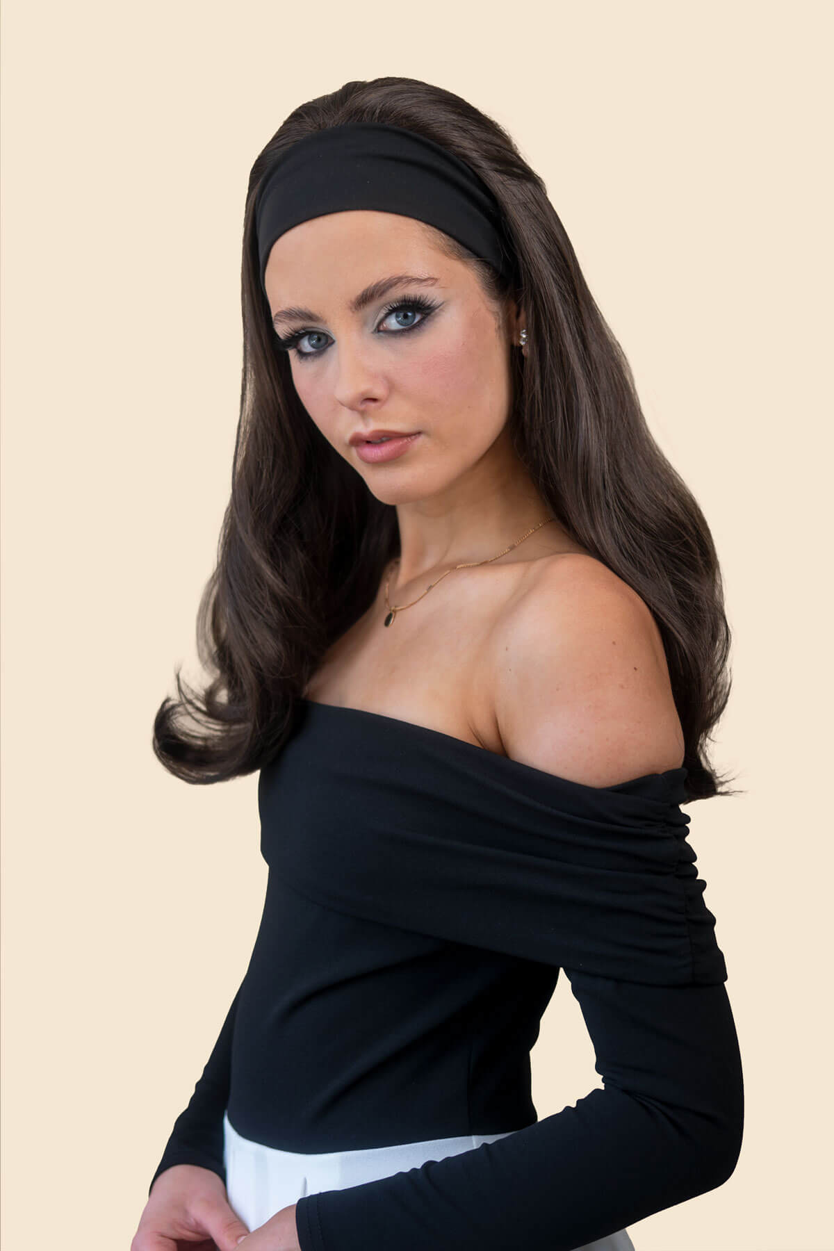 A woman with long dark brown hair wearing a black off-the-shoulder long-sleeve top, a black headband, and a white bottom, posed against a solid light beige background.