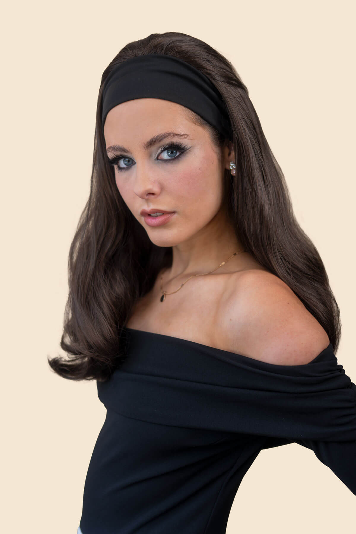 A woman with long dark brown hair wearing a black off-the-shoulder long-sleeve top, a black headband, and a white bottom, posed against a solid light beige background. Long Gently Layered Half Wig with curls towards the tips: Jessica black 1b